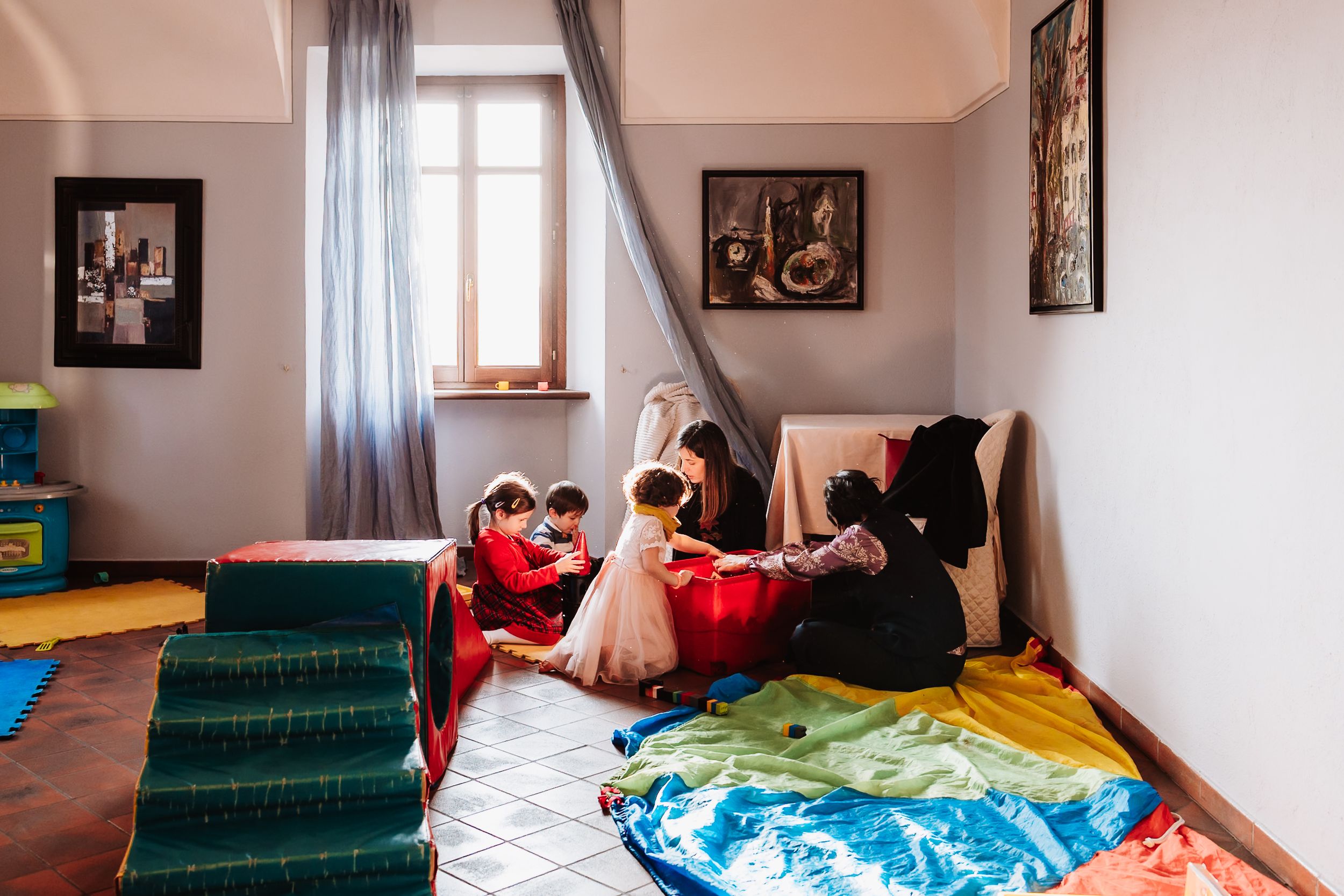 Children playing together in a colorful indoor playroom with soft mats, foam steps, and bright floor cushions near a window.