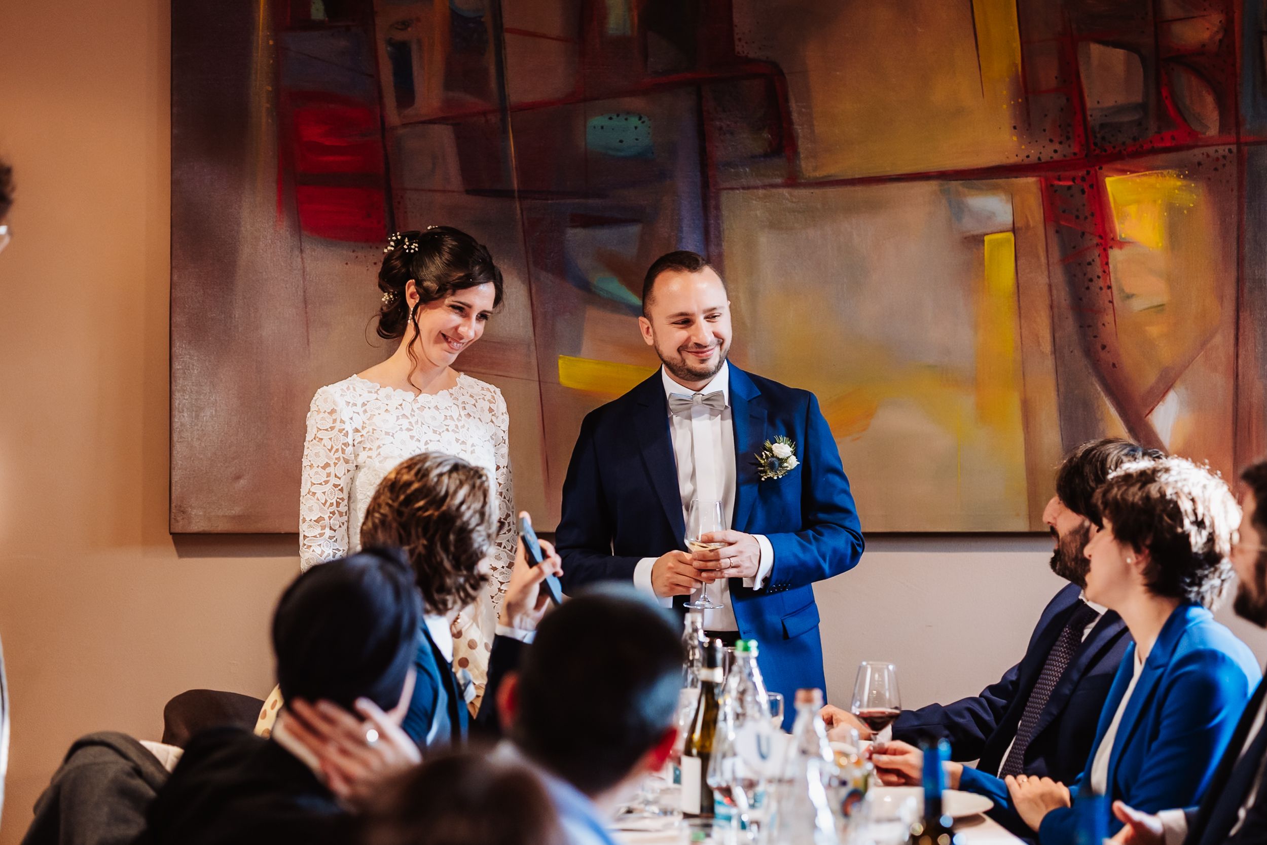 A smiling bride and groom stand together at their wedding reception dinner, engaging with seated guests before colorful artwork.