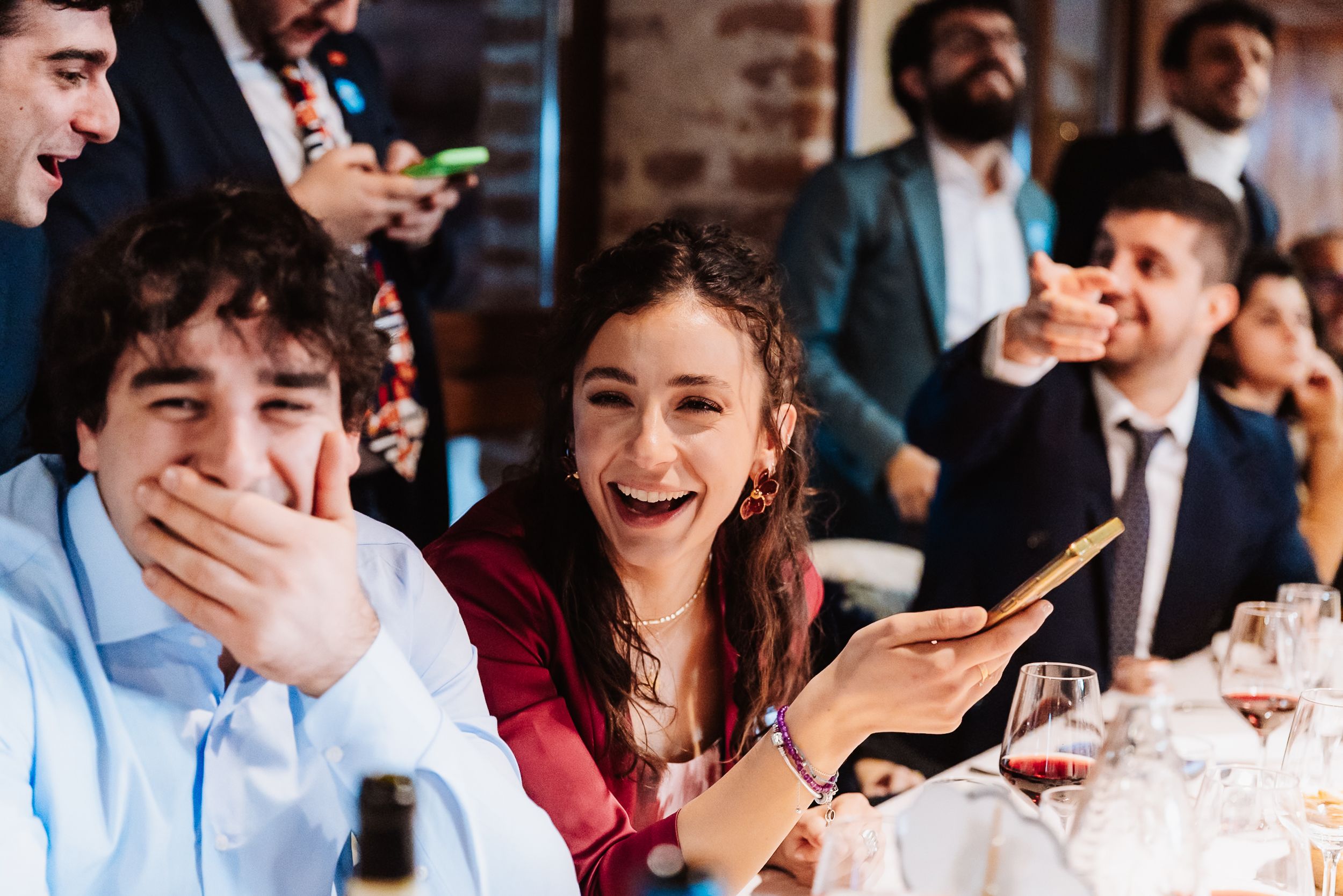 People laughing and smiling at a festive dinner party, with a woman holding a phone and guests enjoying wine.