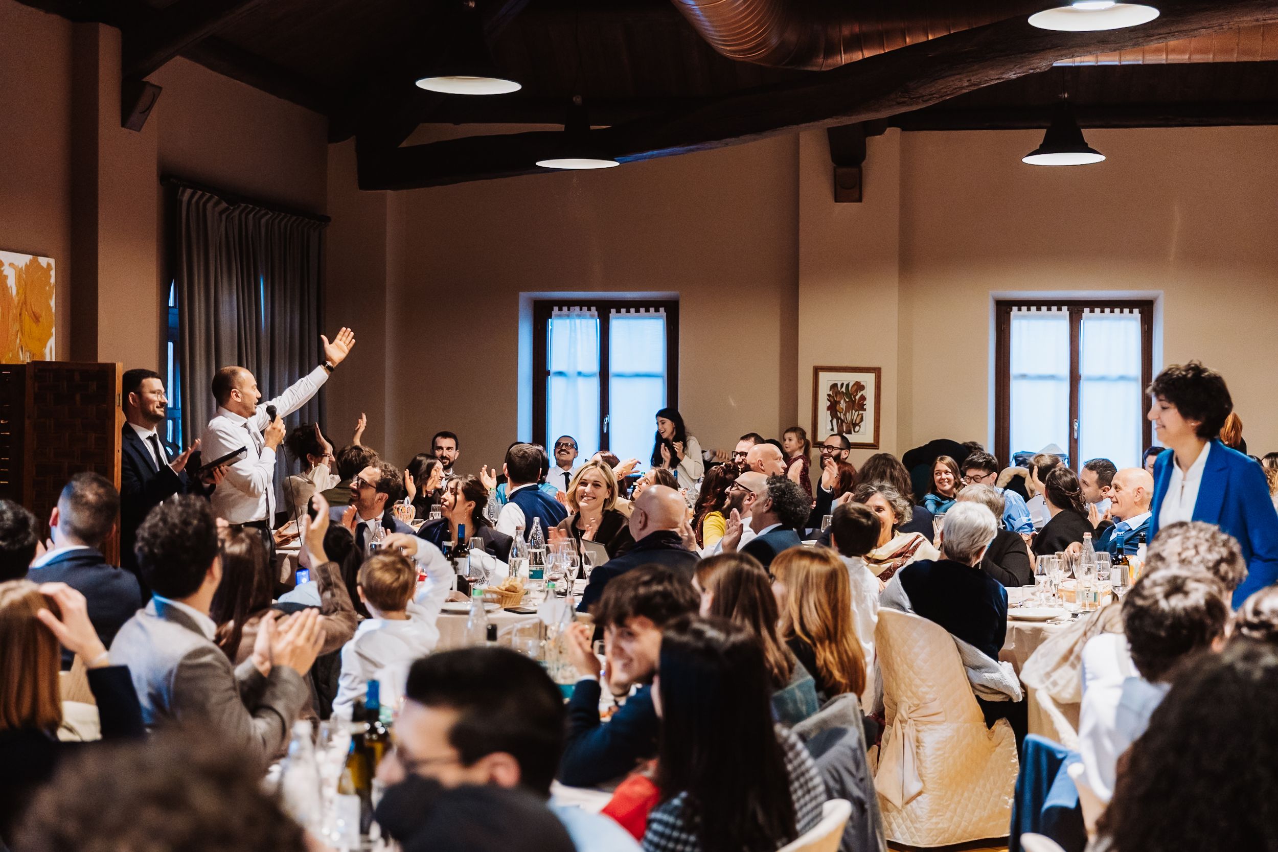 Lively wedding reception in a rustic hall with guests seated at tables and a man raising his hand.