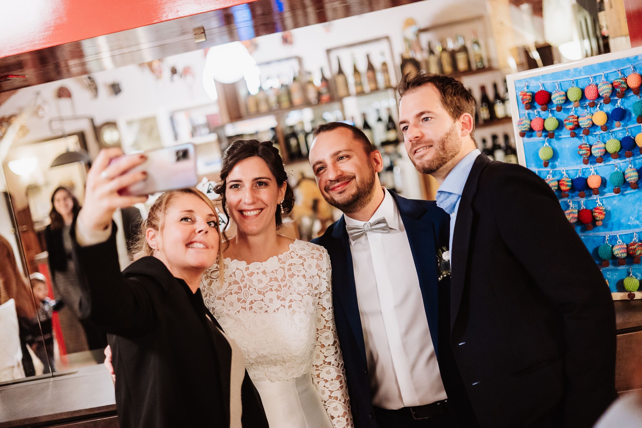 Wedding couple poses for a selfie with guests at a bar venue, bride in white dress, groom in dark suit.