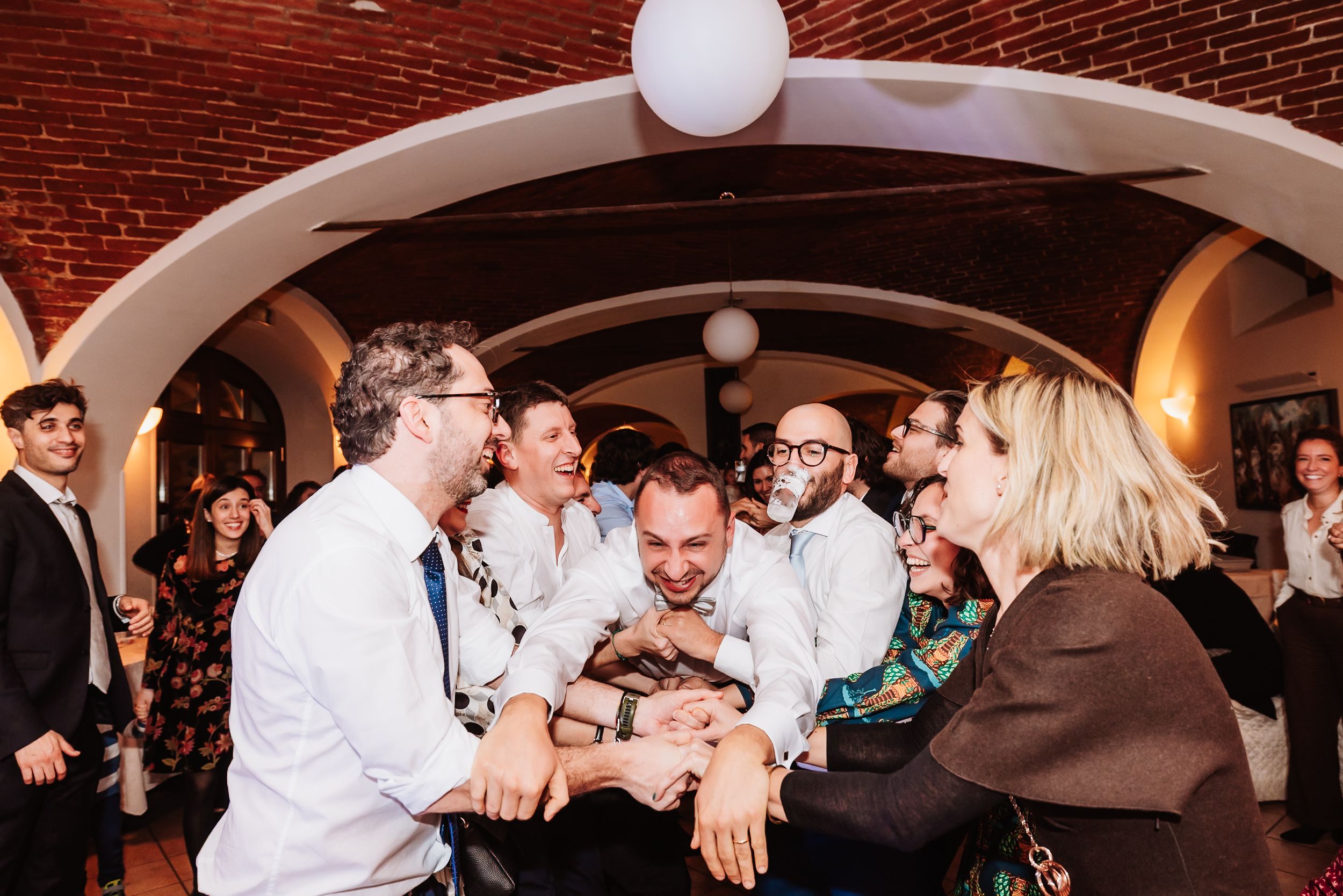 Guests celebrating joyfully at a wedding reception in a brick-arched venue, lifting someone in a lively group moment.