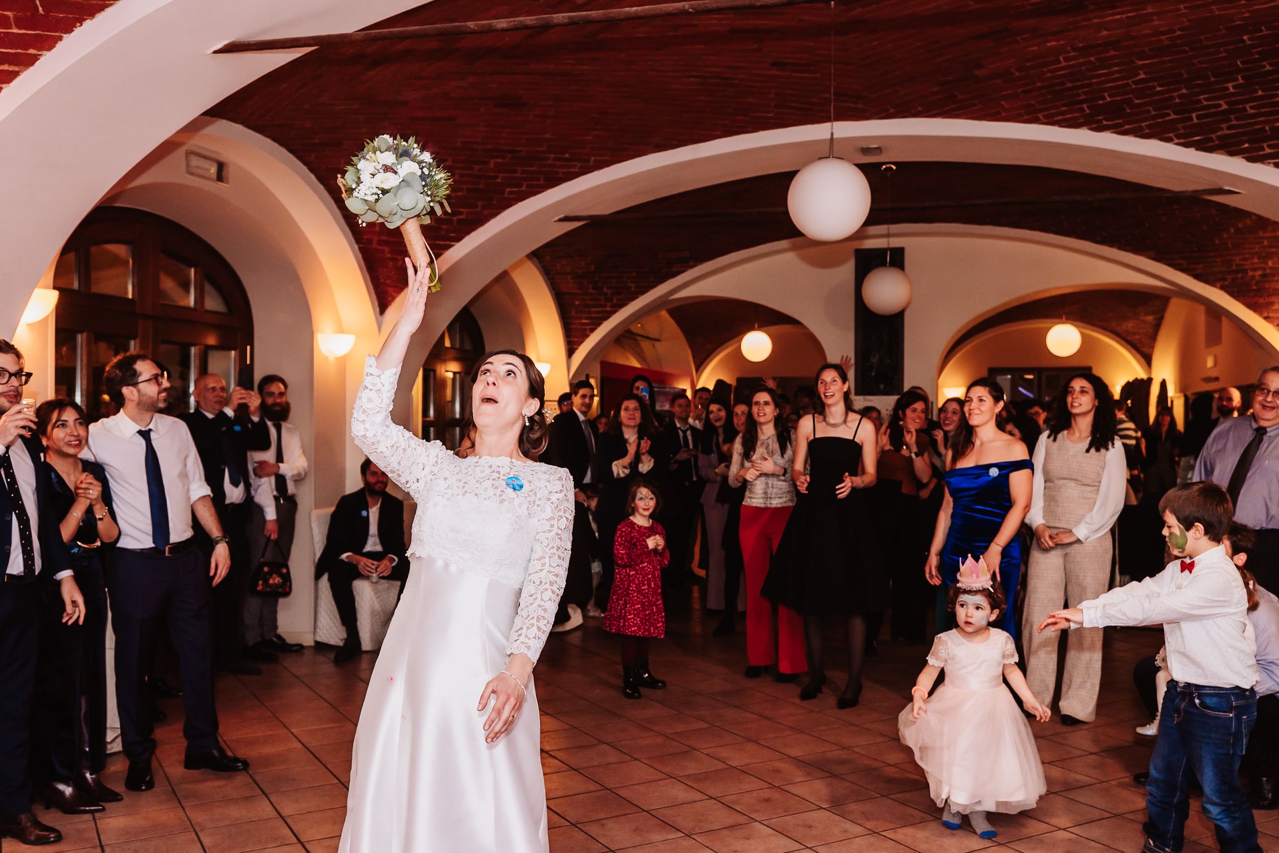 Bride tosses wedding bouquet to guests at elegant indoor reception with arched ceilings.