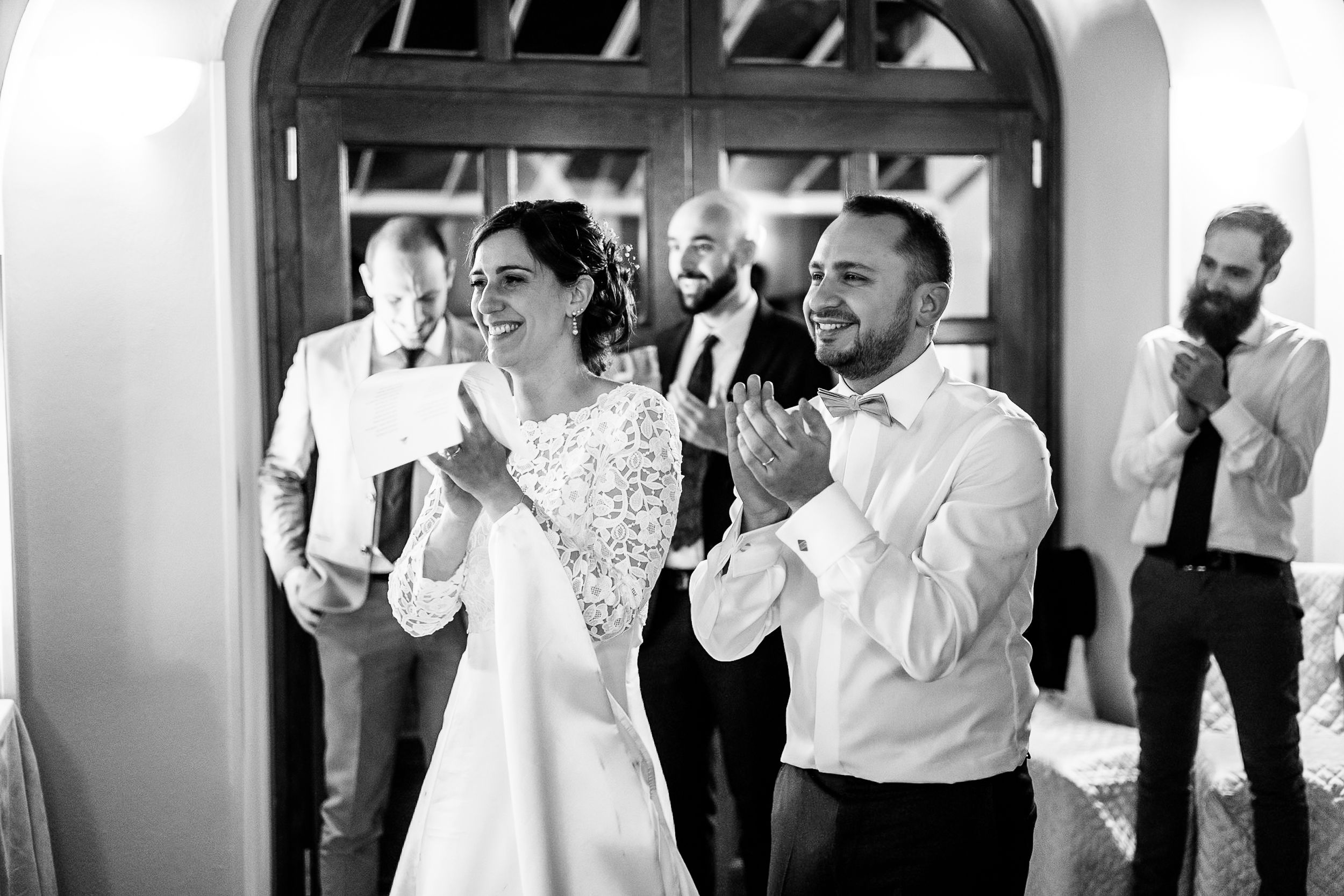 Black and white wedding photo of bride and groom clapping joyfully with guests in elegant venue.