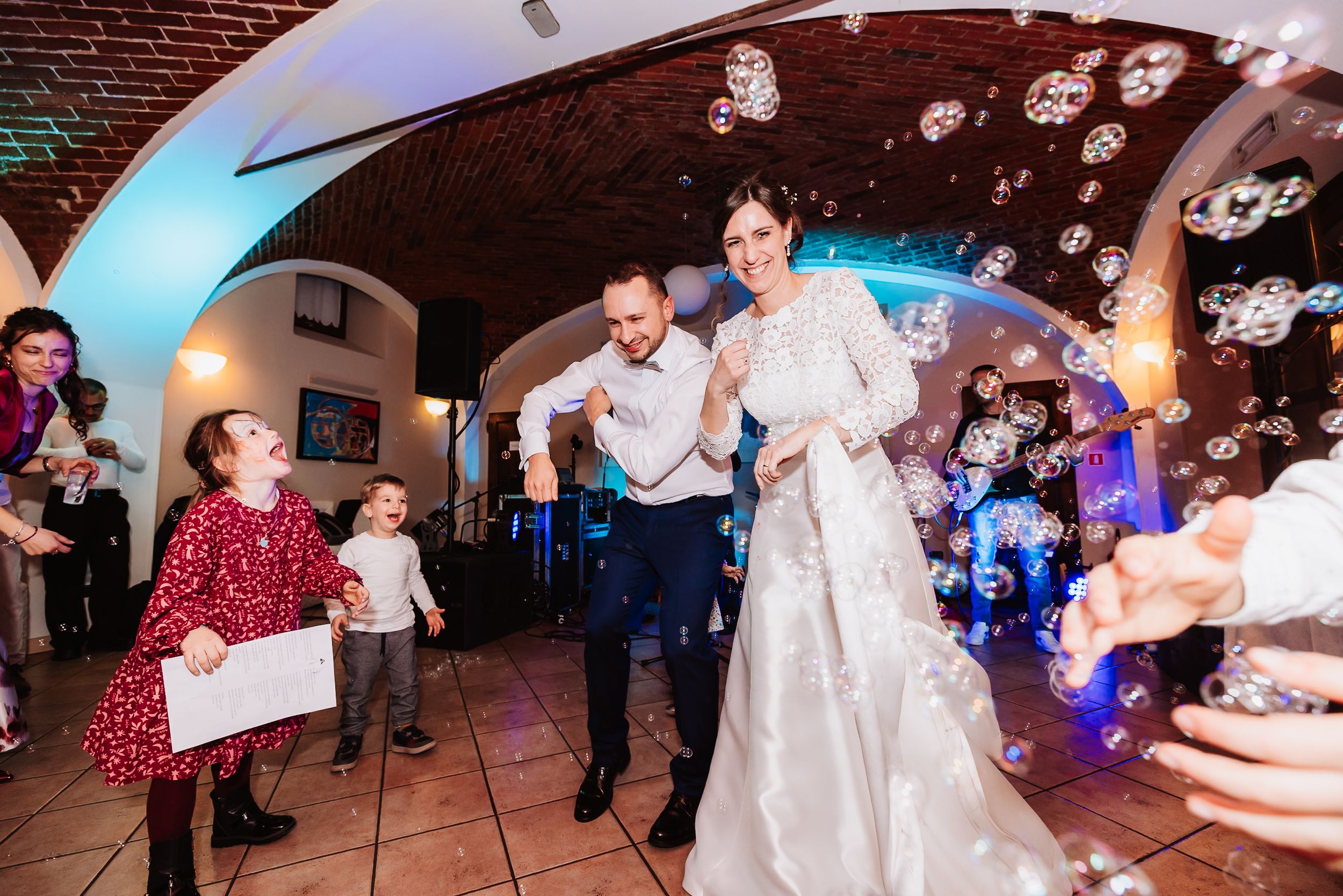 Bride and groom dancing with children surrounded by floating bubbles at an elegant wedding reception venue.
