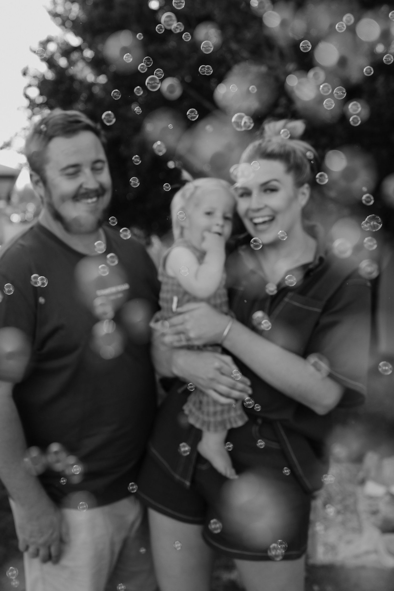 Black and white photo of a happy family with a toddler surrounded by bubbles outdoors near a tree.