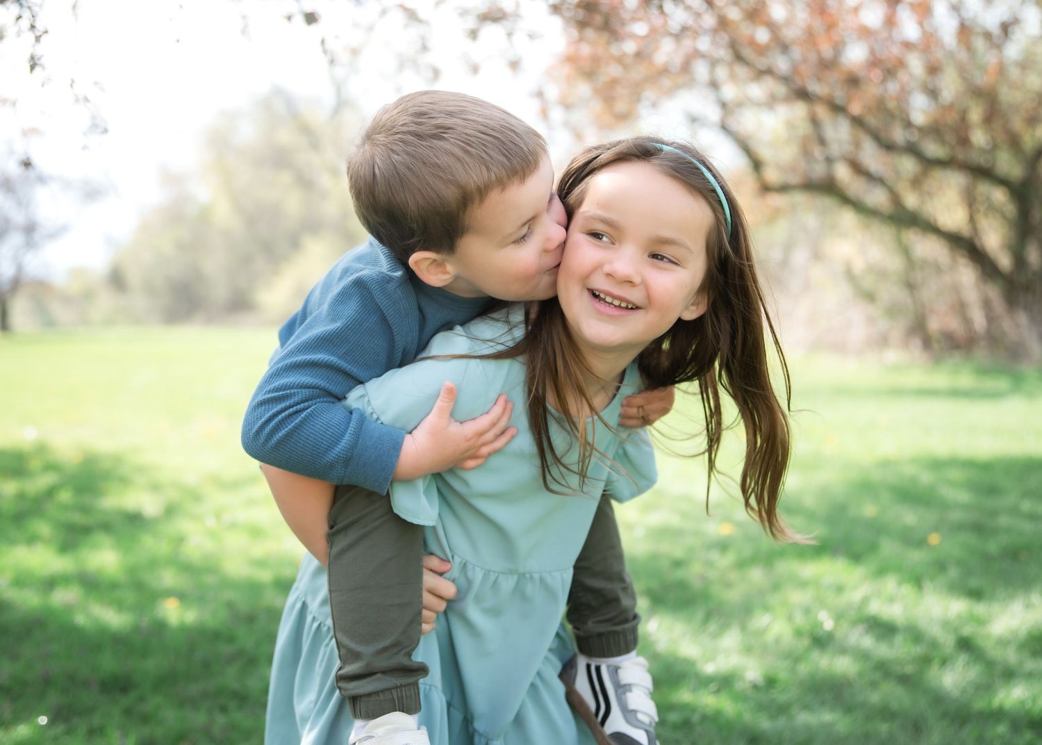 Two young siblings share a sweet moment outdoors, with a boy kissing his smiling sister on the cheek in a green park.
