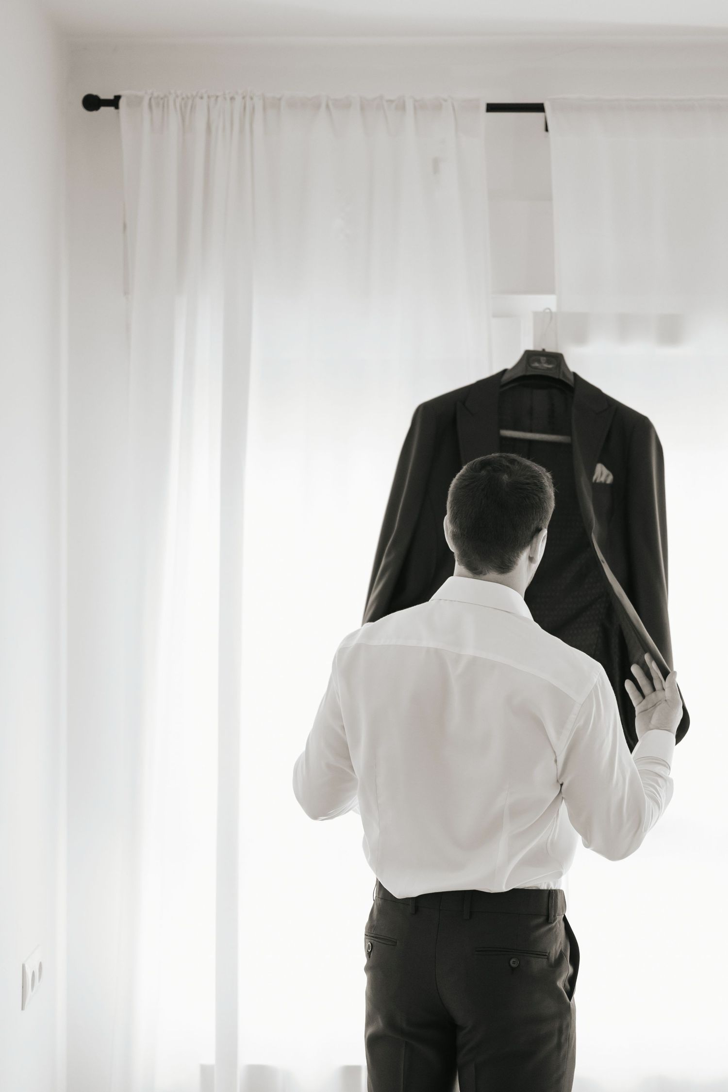 Groom in white shirt reaching for his dark suit jacket hanging by a bright window before his wedding ceremony.