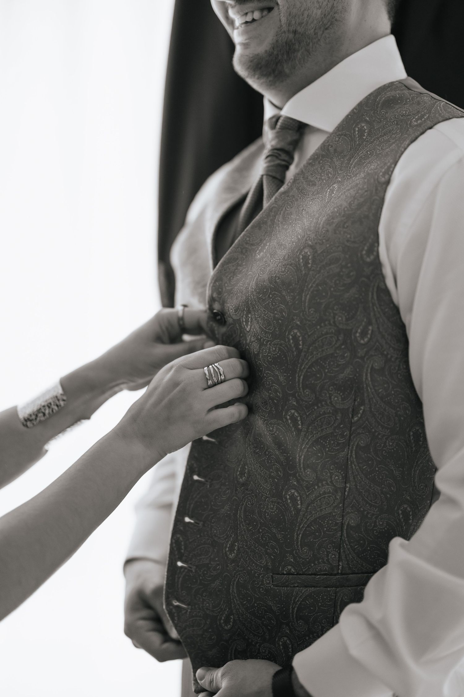 Black and white close-up of hands adjusting a grooms paisley vest on his wedding day.