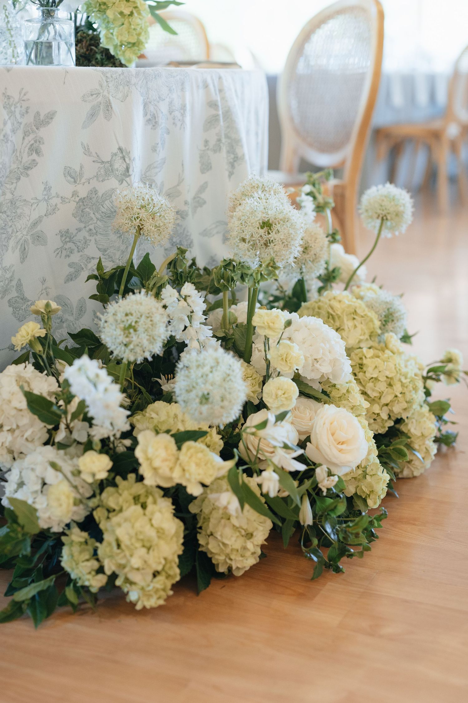 Lush floor arrangement of white roses, hydrangeas, and alliums beside an elegant floral tablecloth at a wedding reception.