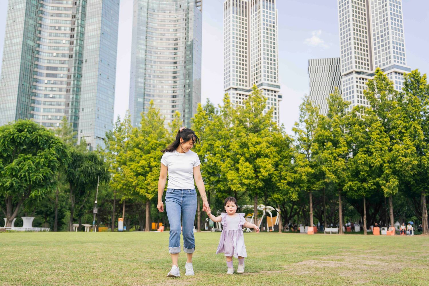 A mother and young daughter hold hands walking in a green urban park with tall skyscrapers in the background.