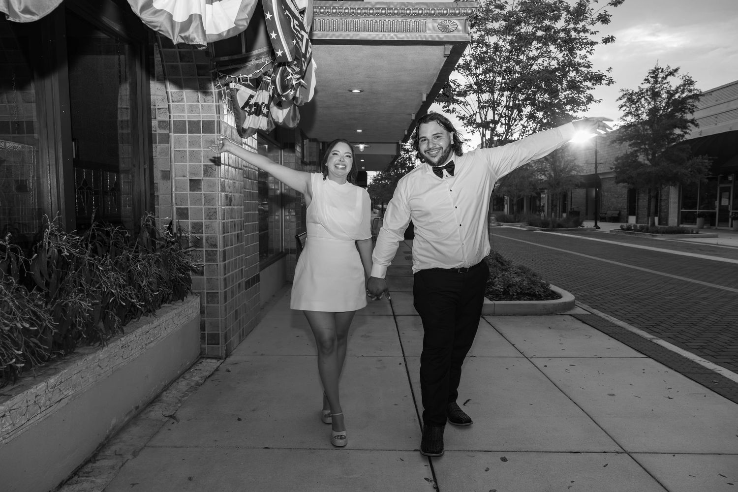 Black and white photo of a joyful couple holding hands with arms raised on a sidewalk outside a brick building.