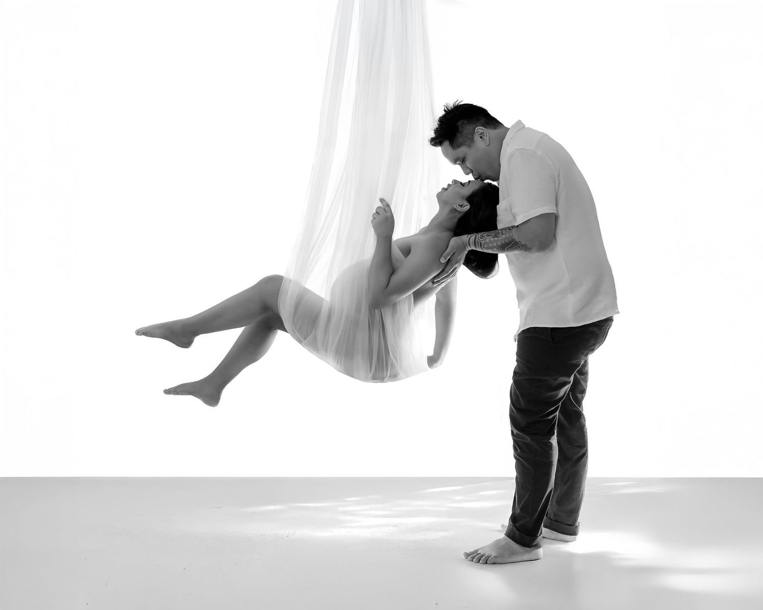 Parents-to-be, pregnant woman hanging from a net, her husband stands his hands on her shoulders leaning over her for a kiss, in studio.