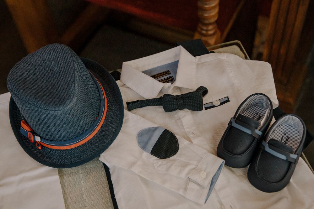 Boys formal outfit flat lay with fedora hat, white dress shirt, bow tie, socks, and black leather shoes.
