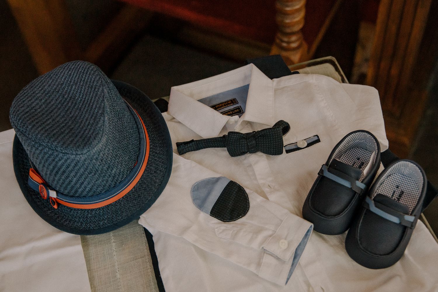 Boys formal outfit flat lay with fedora hat, white dress shirt, bow tie, socks, and black leather shoes.