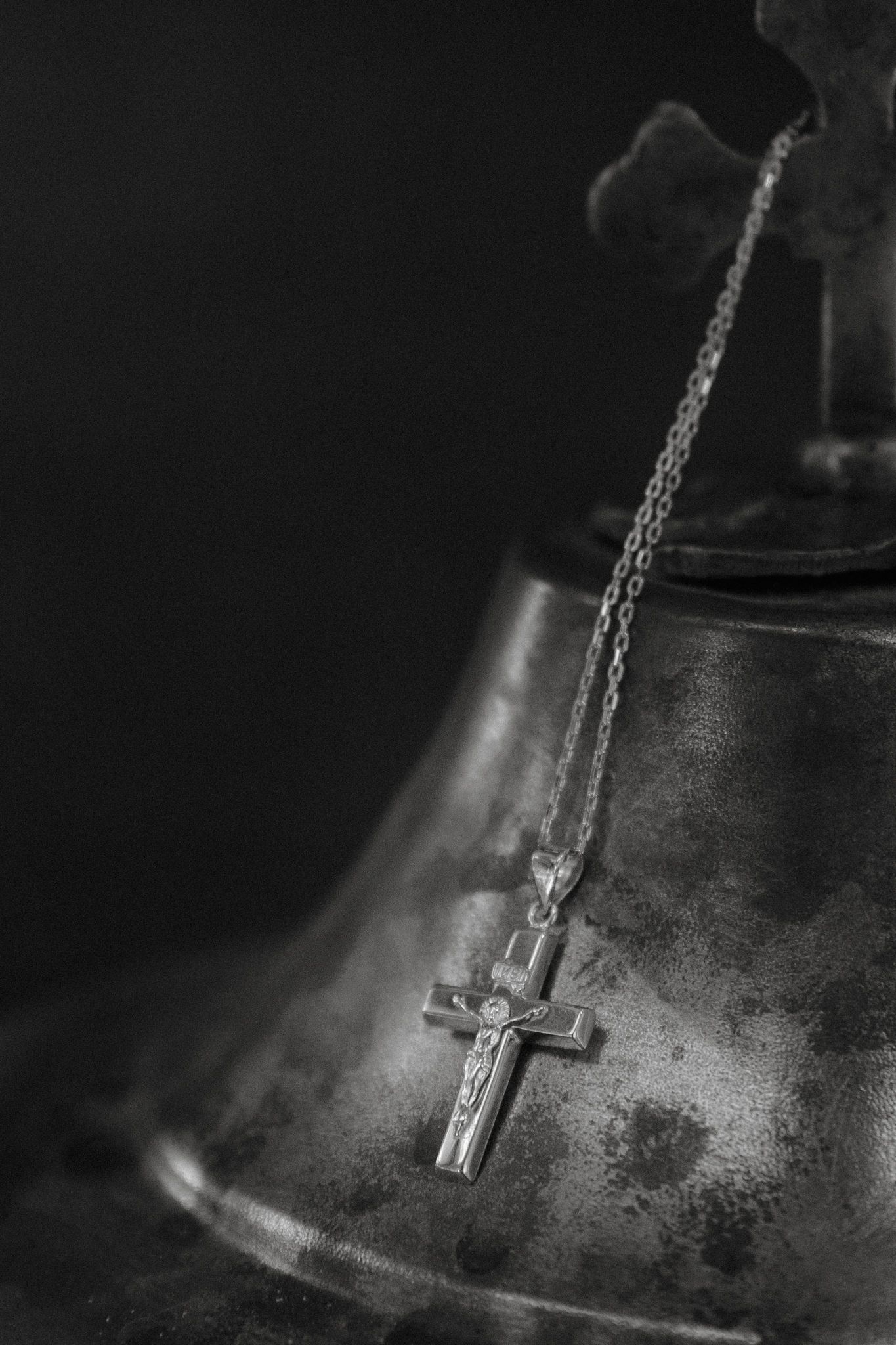 Black and white photo of a crucifix necklace draped over an antique metal bell with a cross on top.