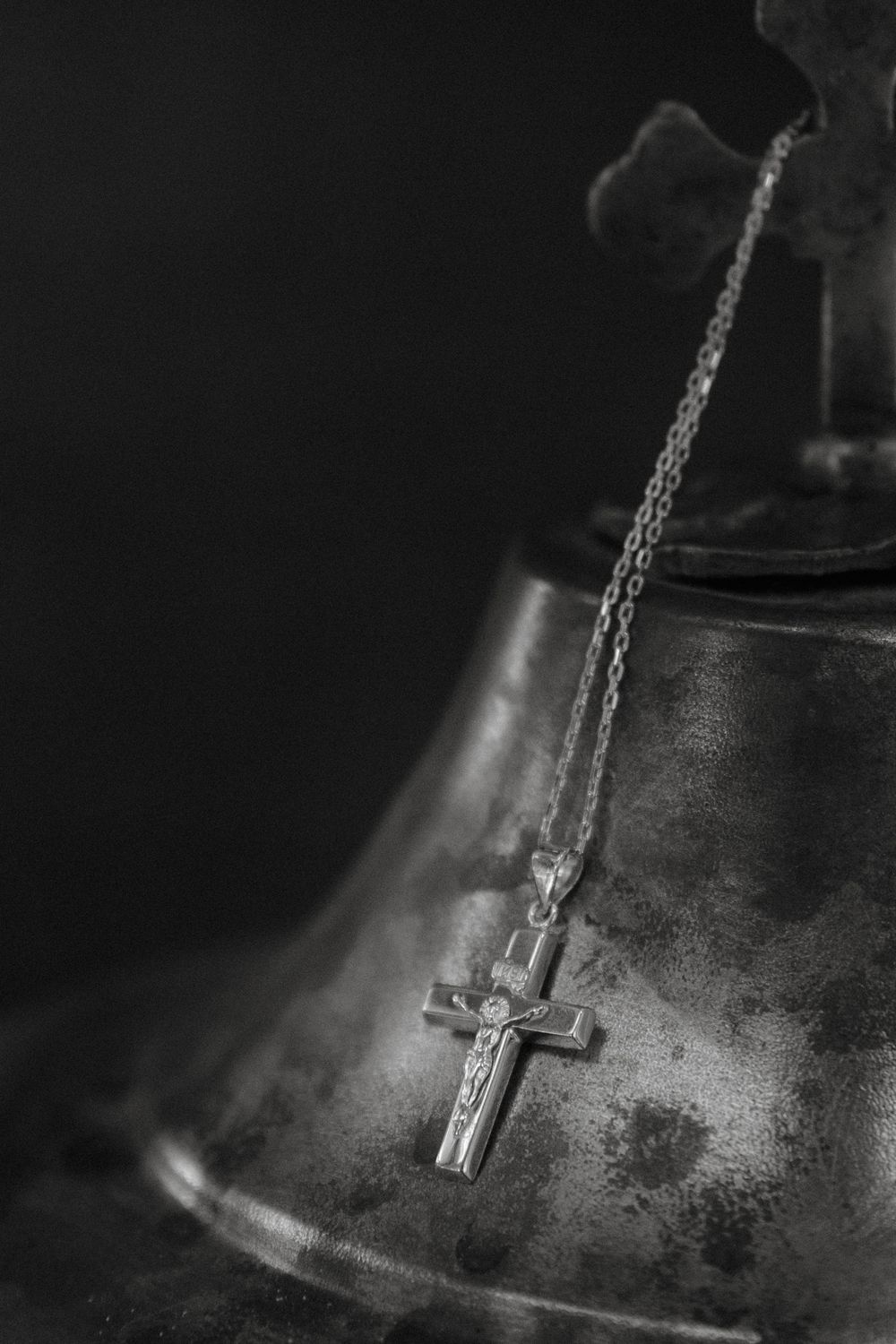 Black and white photo of a crucifix necklace draped over an antique metal bell with a cross on top.