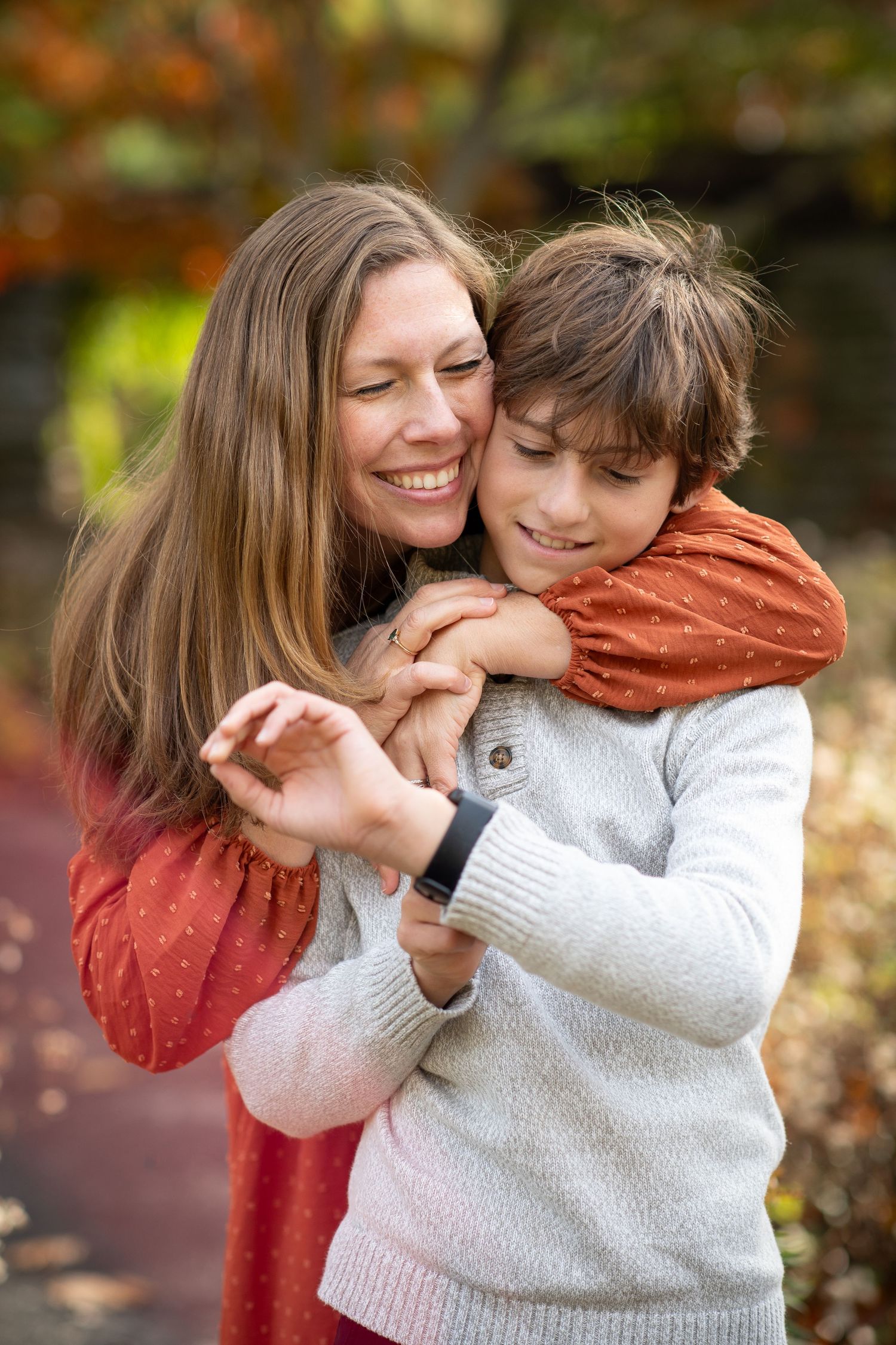 A smiling woman in a red dress hugs a young boy in a white sweater outdoors in an autumn park setting.