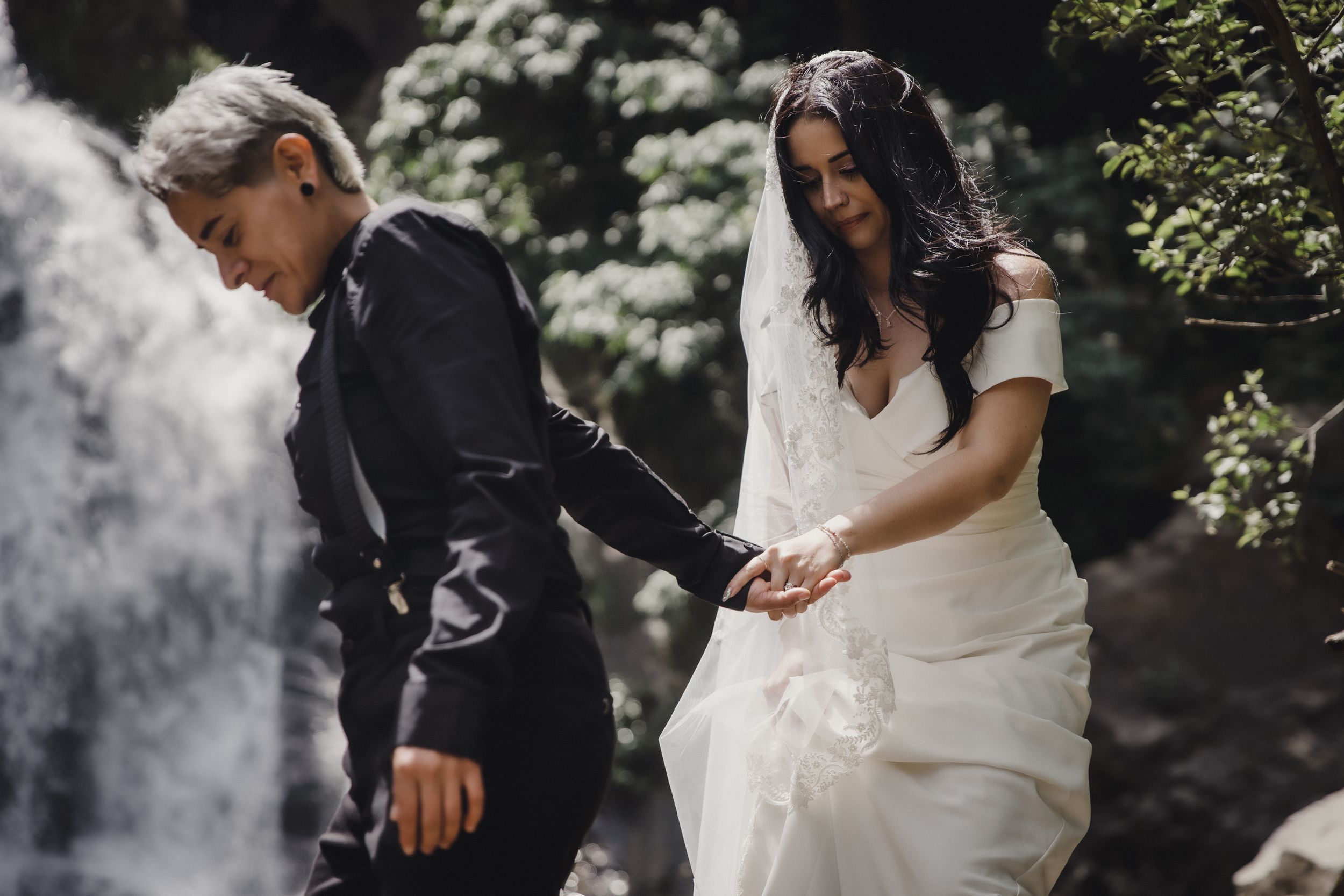 Two brides in wedding attire hold hands near a waterfall, one in a white gown and one in a black suit.