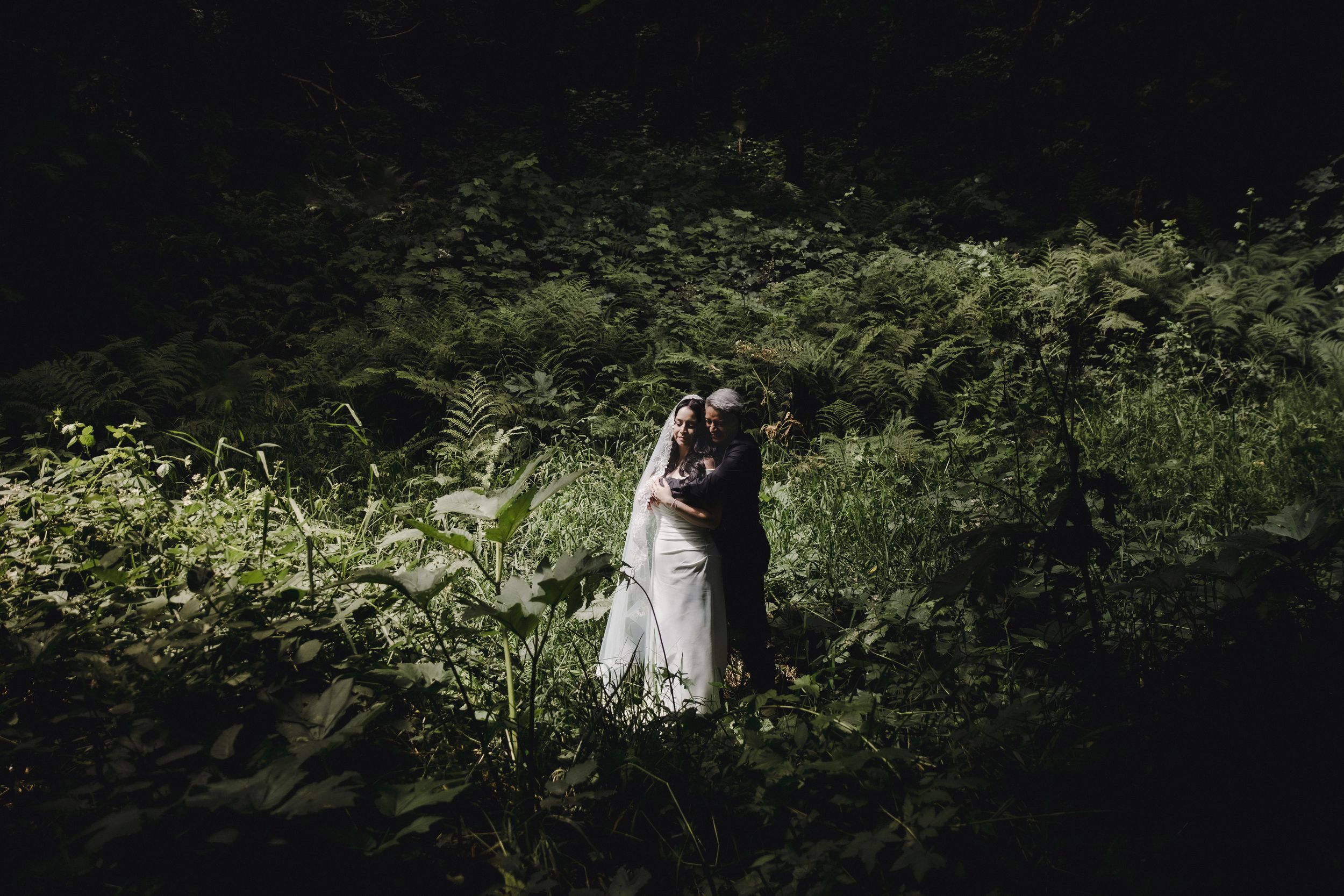 A bride and groom embrace romantically in a lush green forest setting during their outdoor wedding ceremony.