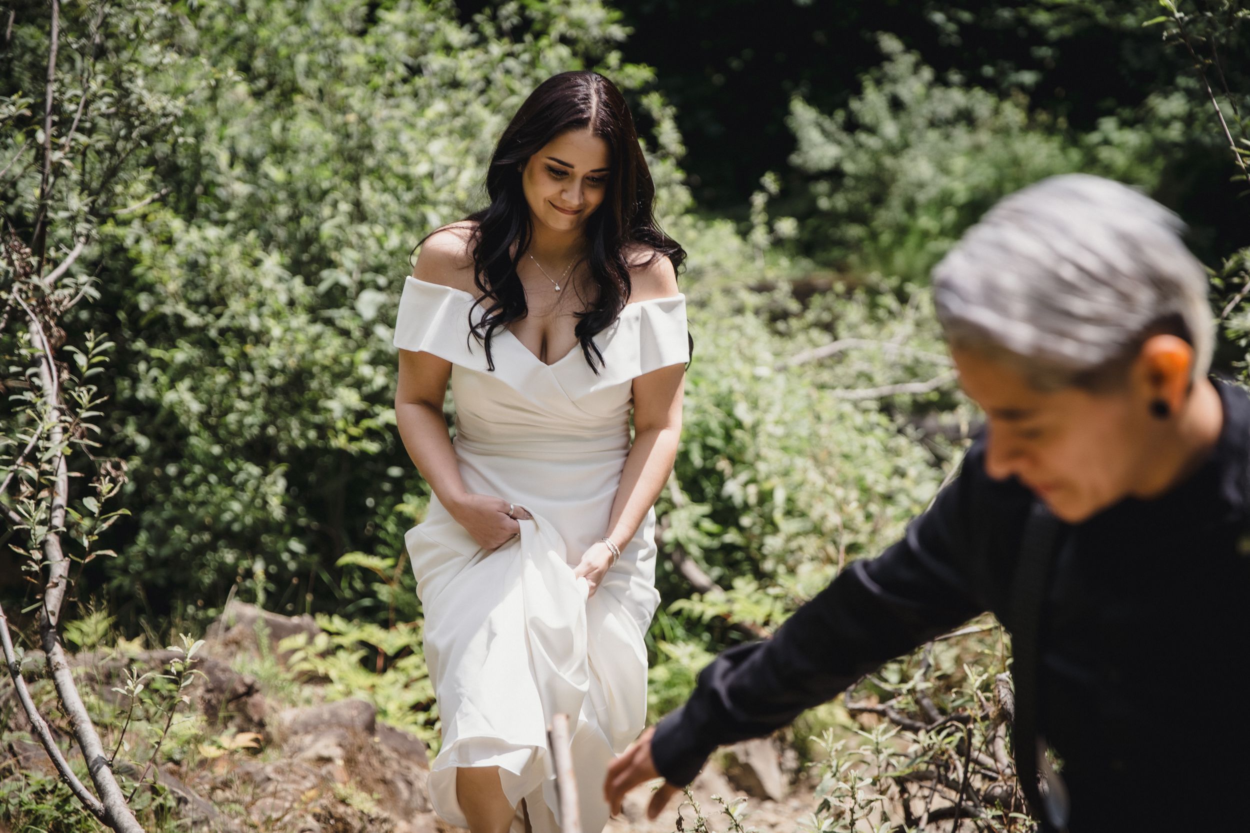 Bride in white off-shoulder gown hiking through forest with helper on her wedding day.