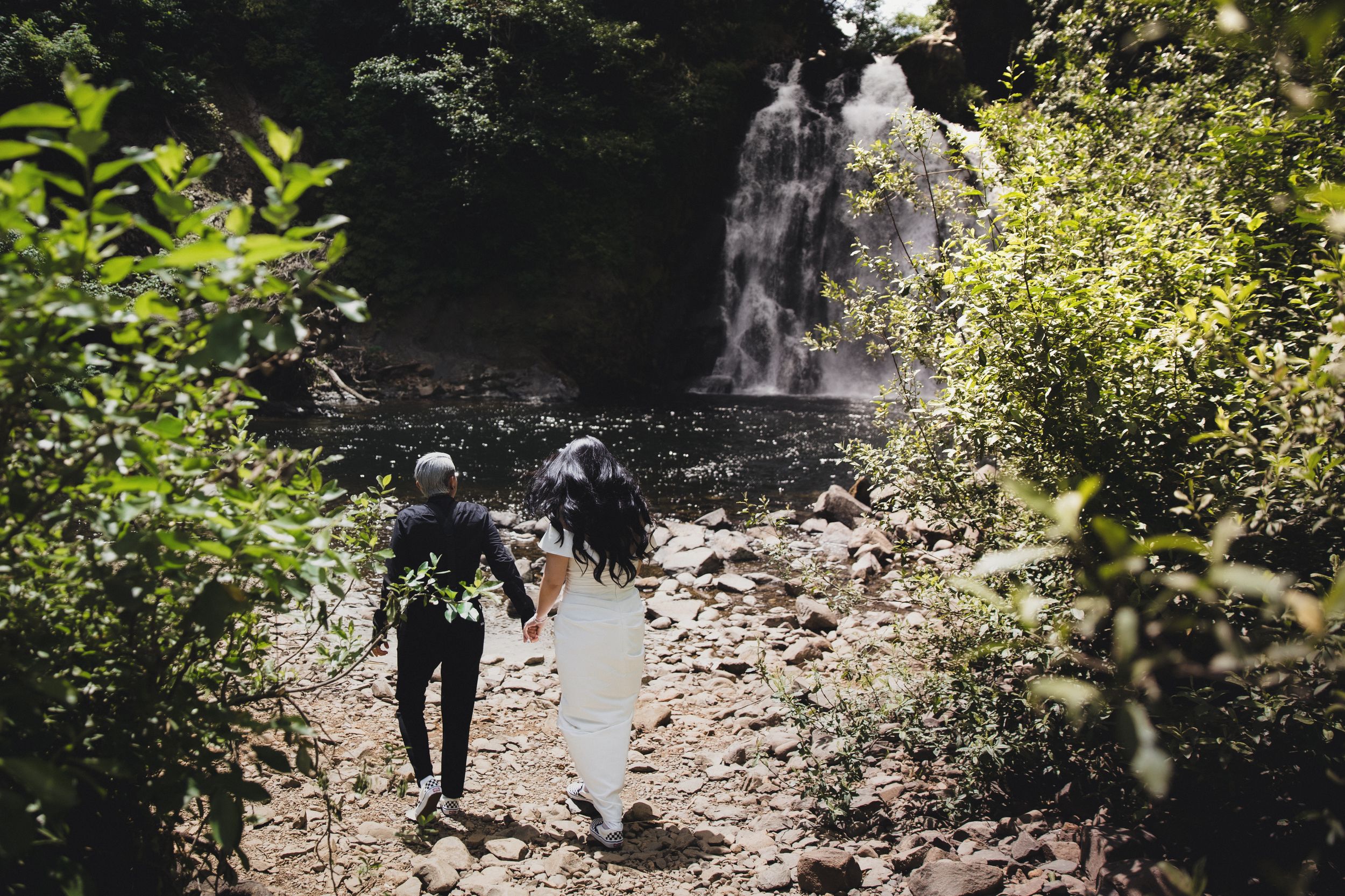 Couple holding hands walking toward a lush tropical waterfall surrounded by green foliage on a rocky path.