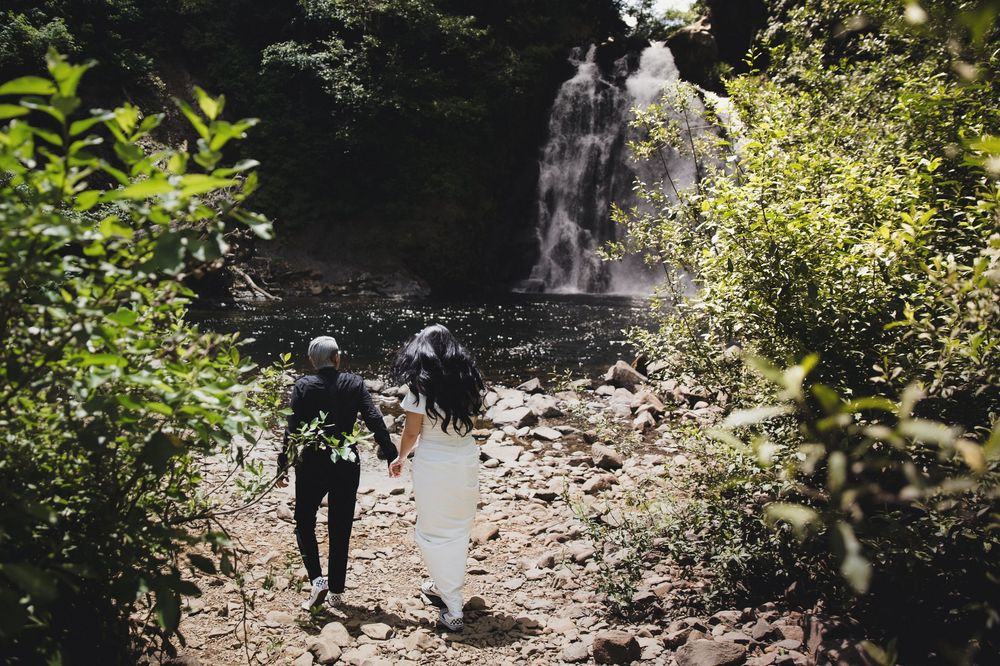 Couple holding hands walking toward a lush tropical waterfall surrounded by green foliage on a rocky path.