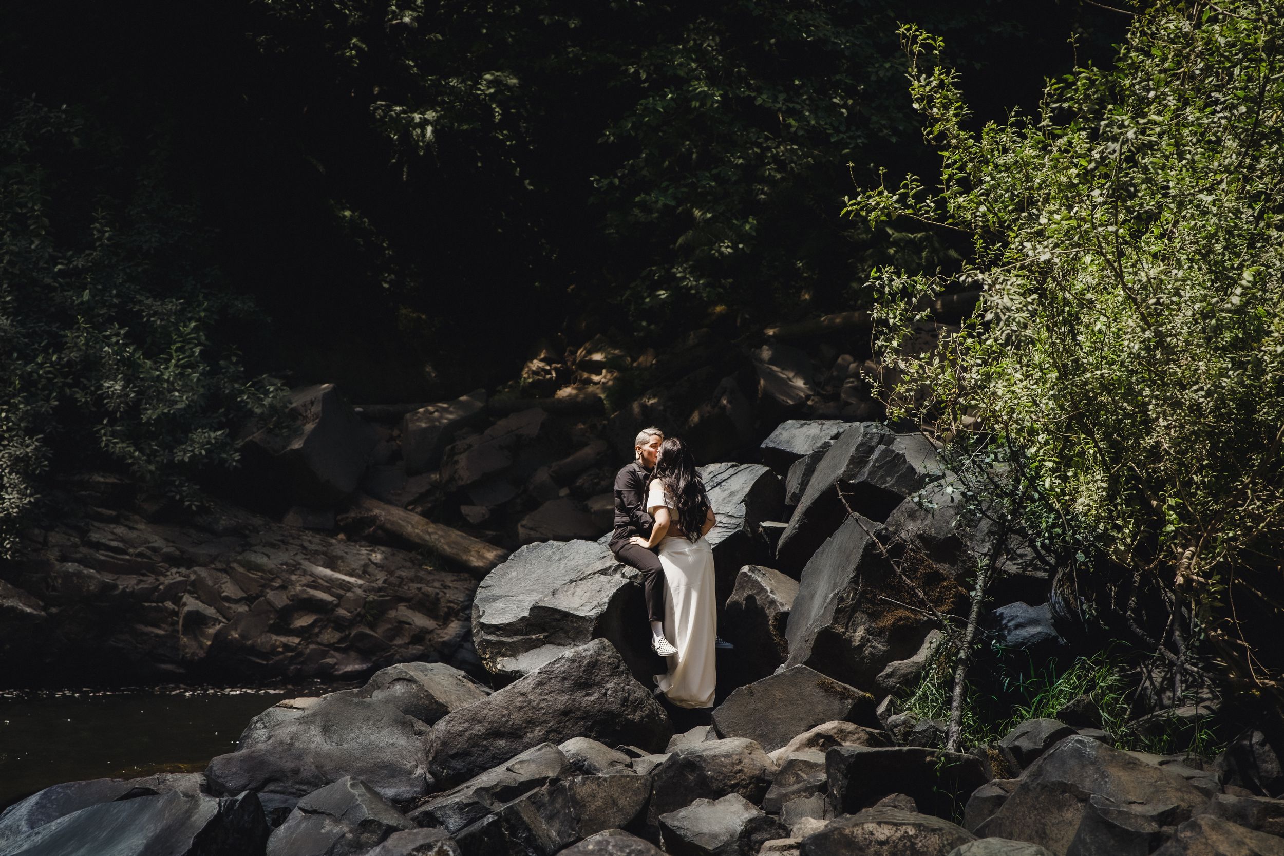 Couple embracing on rocky riverbank surrounded by lush green trees in a romantic outdoor elopement setting.