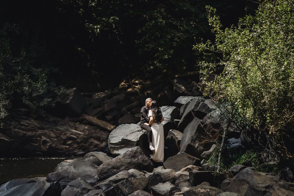 Couple embracing on rocky riverbank surrounded by lush green trees in a romantic outdoor elopement setting.