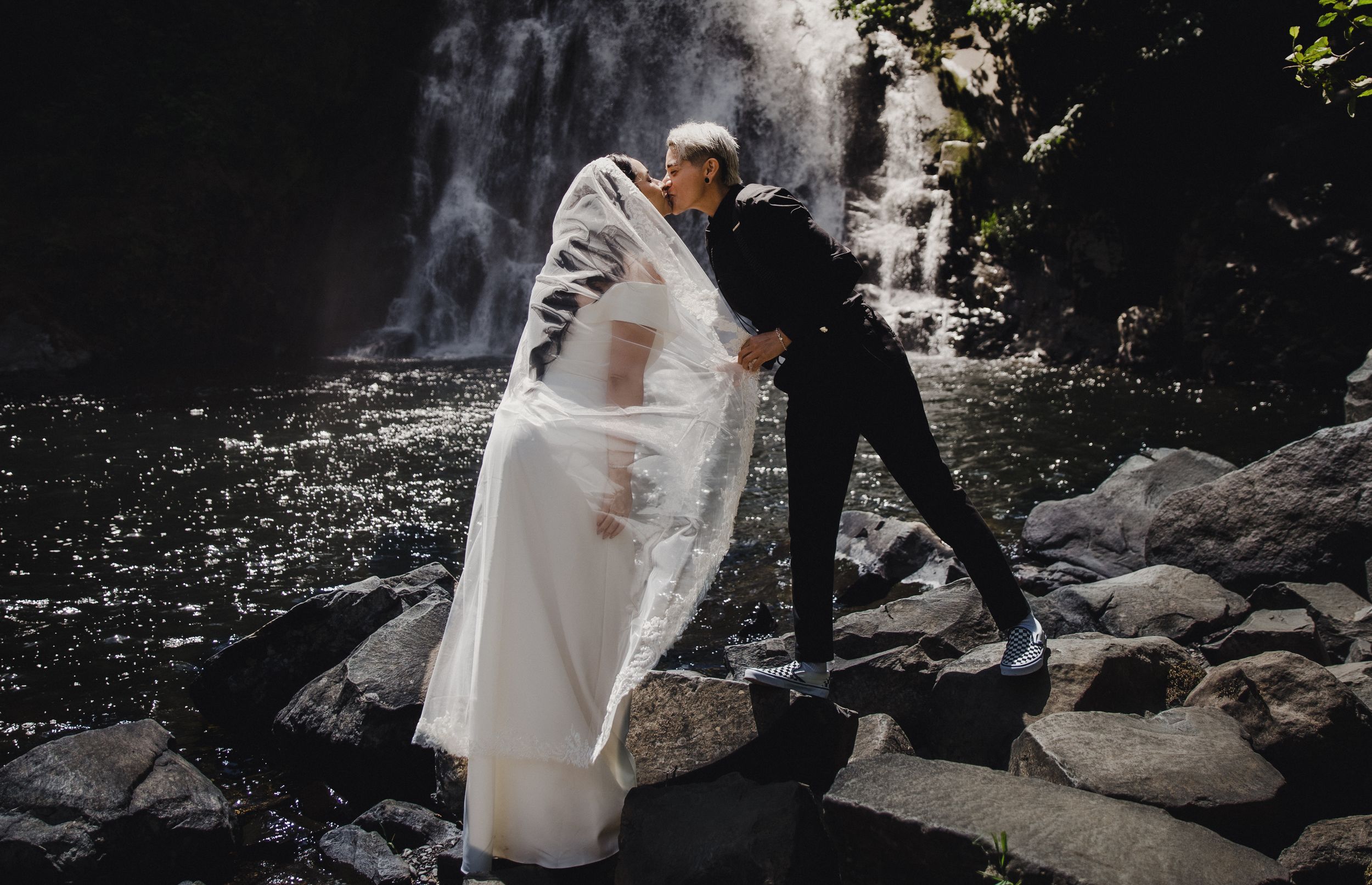 A bride in a flowing white gown and veil kisses her partner near a stunning waterfall surrounded by rocks.