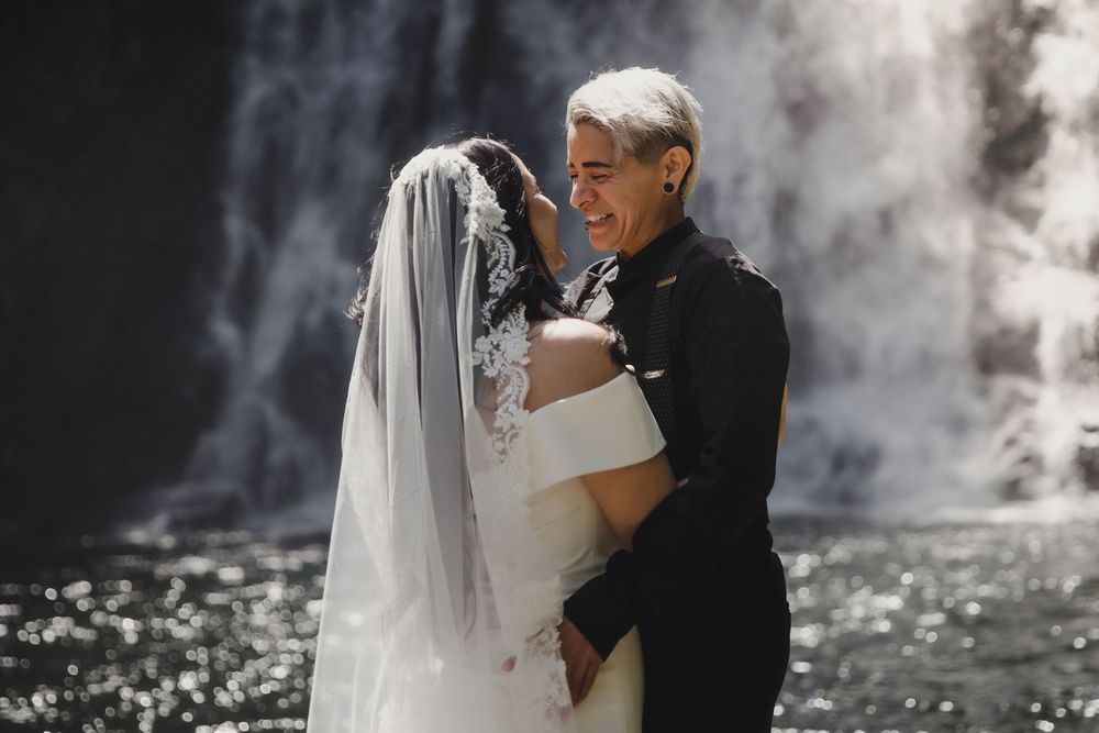 Bride and groom embrace laughing in front of a waterfall, bride in white gown and veil, groom in black suit.