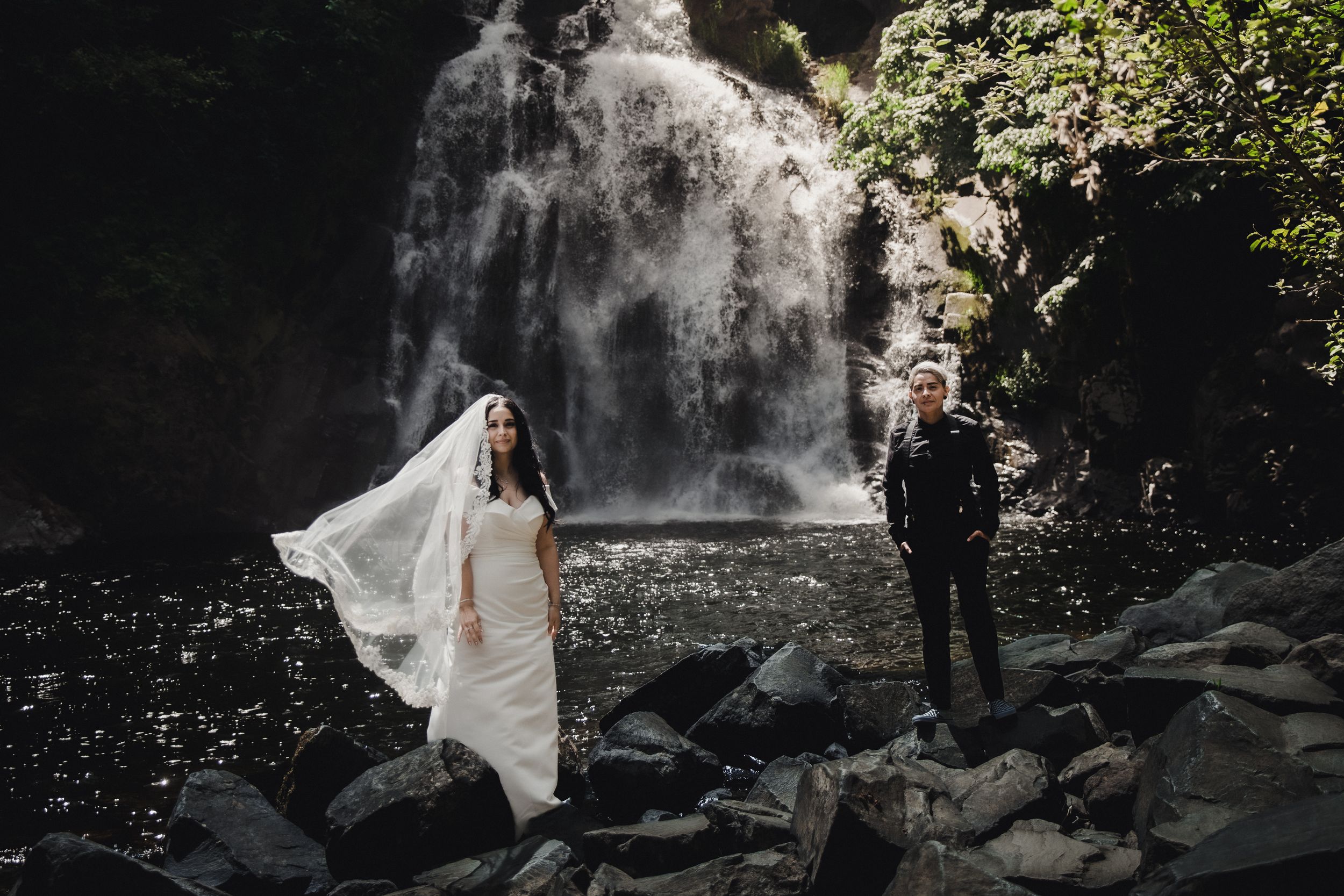 Bride and groom stand on rocky riverbank before stunning waterfall, bridal veil flowing in wind.