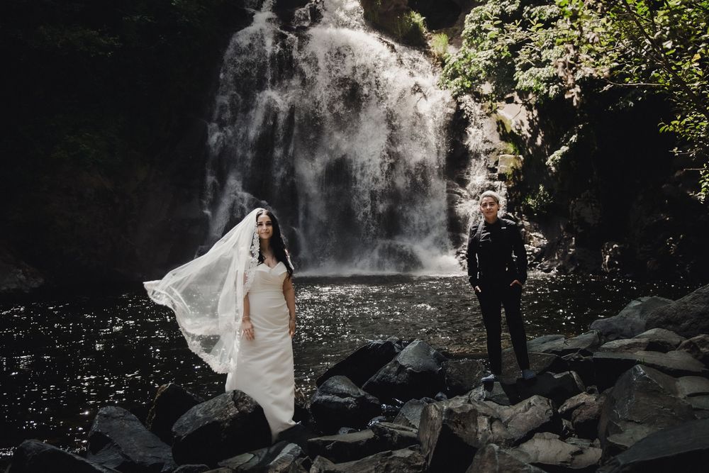 Bride and groom stand on rocky riverbank before stunning waterfall, bridal veil flowing in wind.