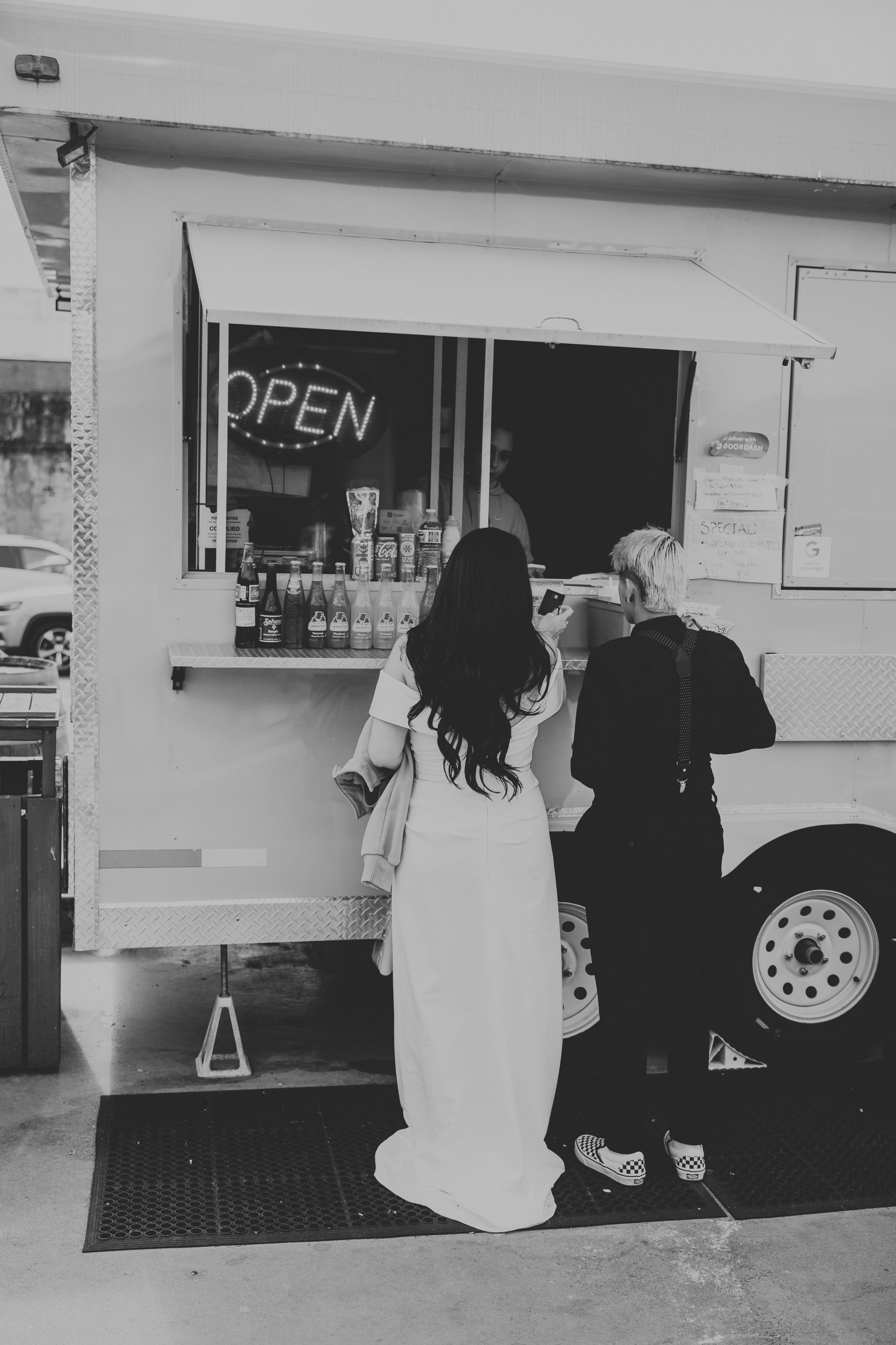 Black and white photo of a bride in a white gown ordering at an open food truck with a companion.