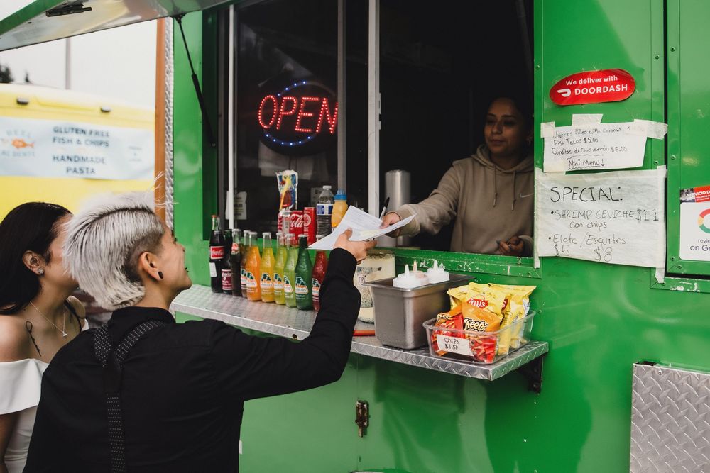 Customers ordering food at a green food truck with an open sign and special menu board displayed.