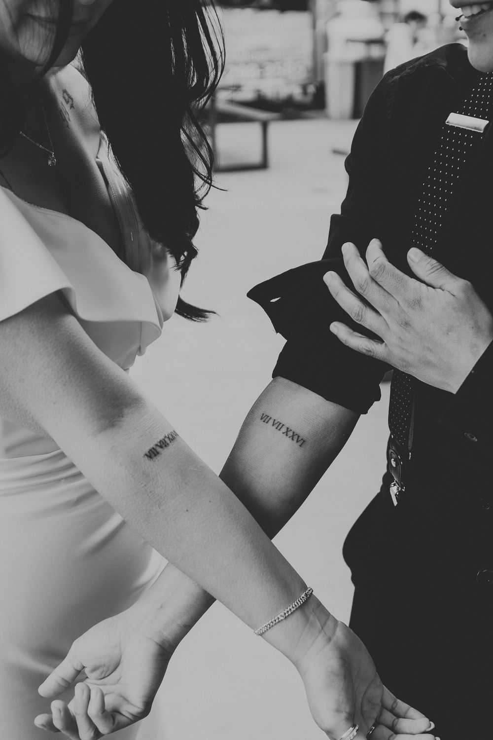 Black and white photo of a couple holding hands showing matching Roman numeral tattoos on their forearms.