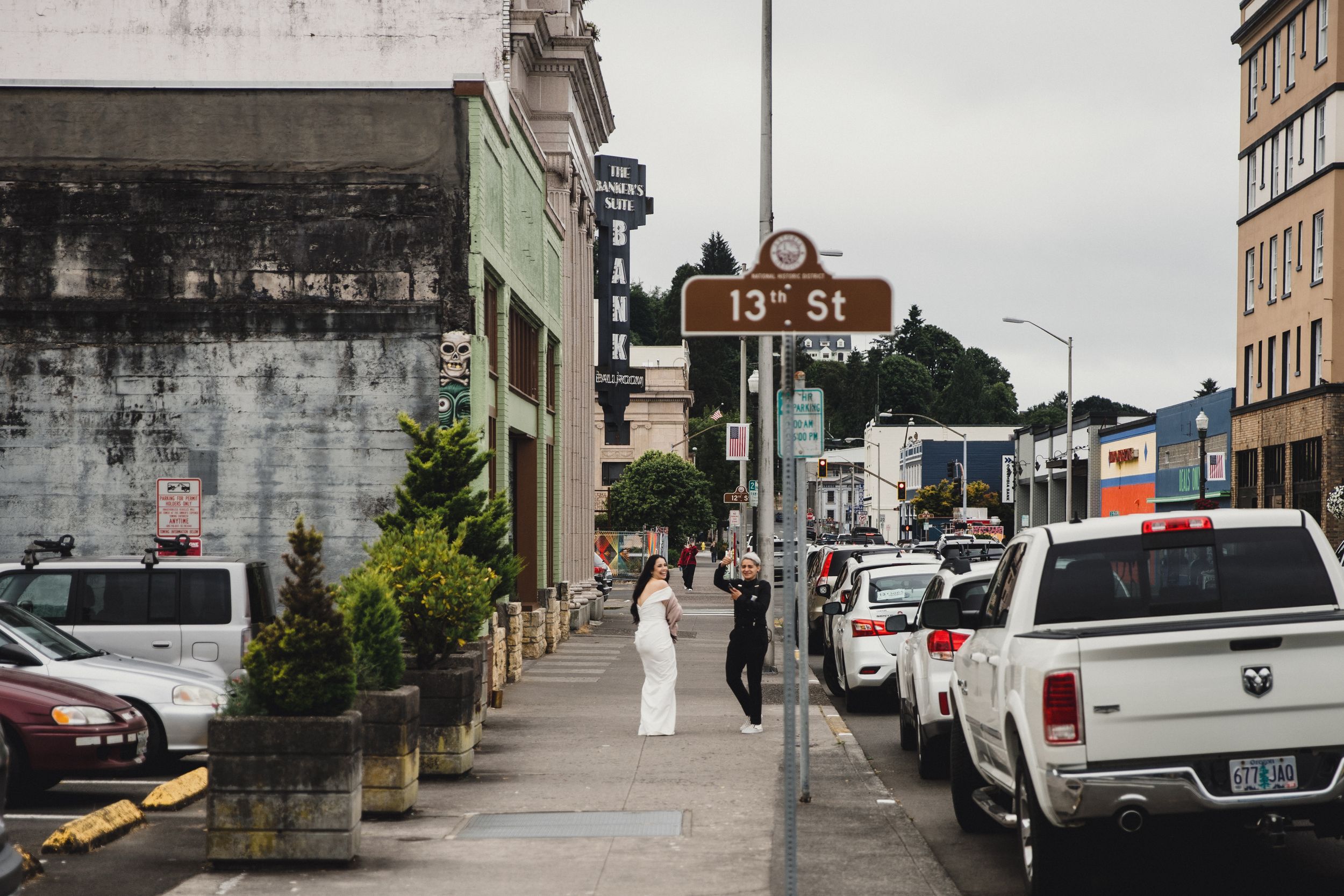 Bride in white dress walks on 13th St sidewalk in urban setting with parked cars and historic buildings on cloudy day.