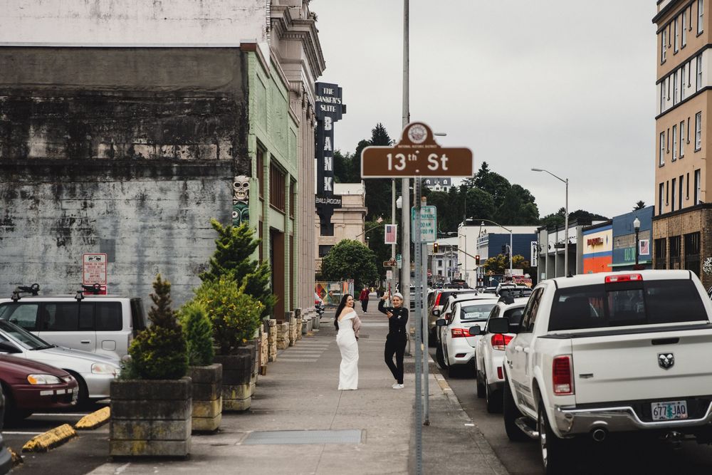 Bride in white dress walks on 13th St sidewalk in urban setting with parked cars and historic buildings on cloudy day.