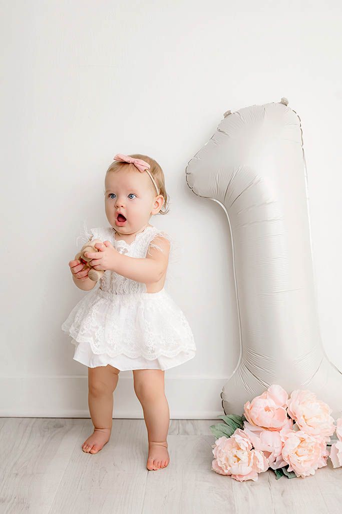 Adorable baby girl in white lace dress stands next to a number 1 balloon and pink flowers for her first birthday.