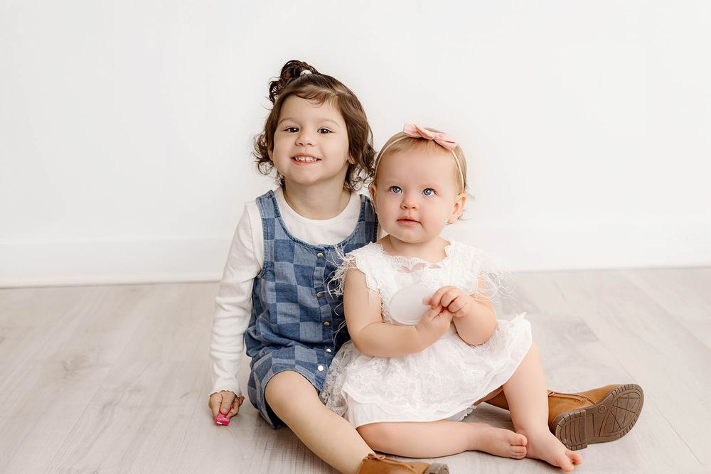 Two young sisters sitting together on a light wood floor, smiling, wearing matching white and blue outfits.
