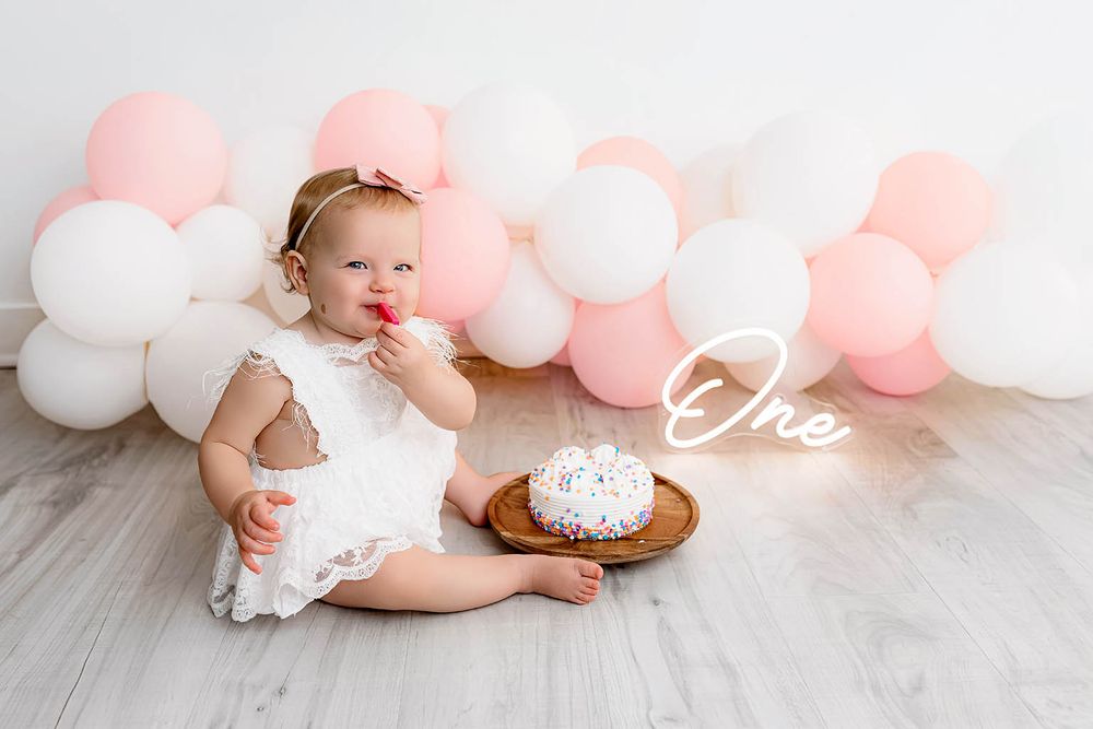 Baby girl in white dress celebrating first birthday with cake and pink and white balloon garland backdrop.