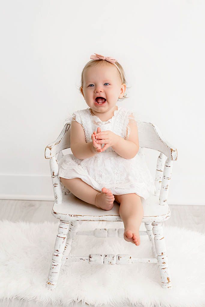 Happy baby girl in white lace dress and pink bow sits clapping on a rustic white chair during a studio portrait session.
