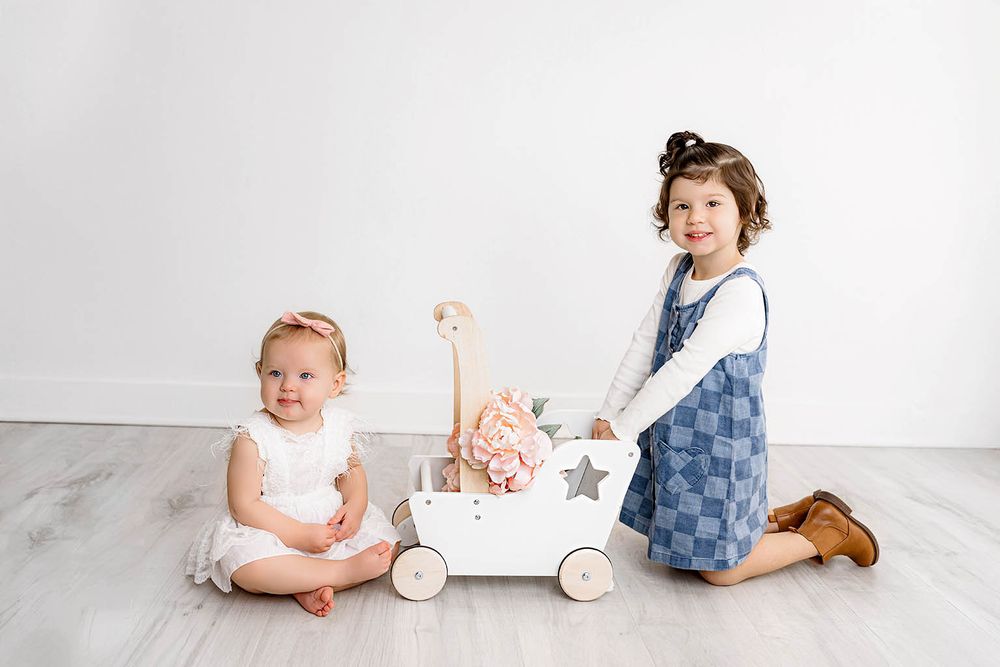 Two young girls pose with a white wooden toy cart featuring stars in a bright, minimalist studio setting.