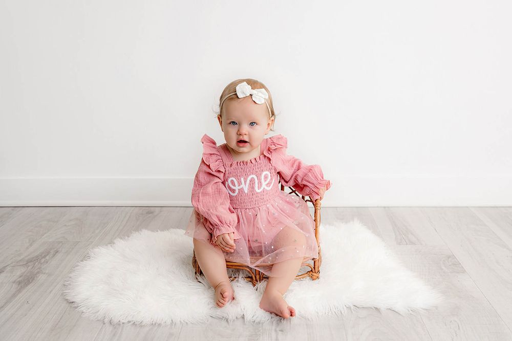 Baby girl in pink 'one' dress sits on fur rug for first birthday photo session.
