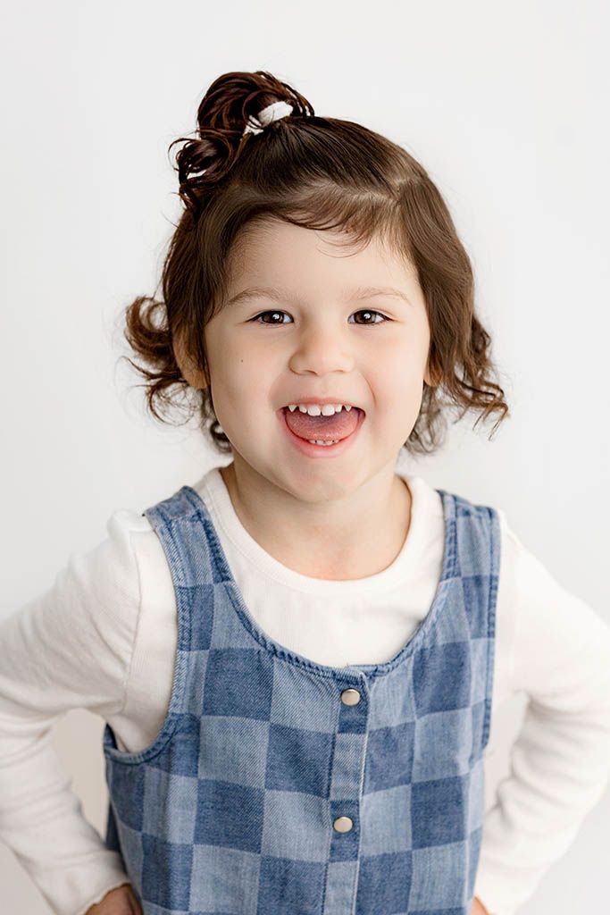 Smiling toddler girl in checkered denim jumper poses for studio portrait photo.