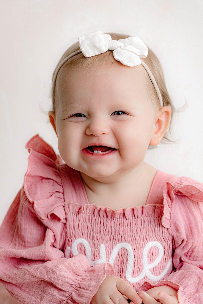 Adorable baby girl in pink \"one\" dress with white bow headband smiling for first birthday portrait.