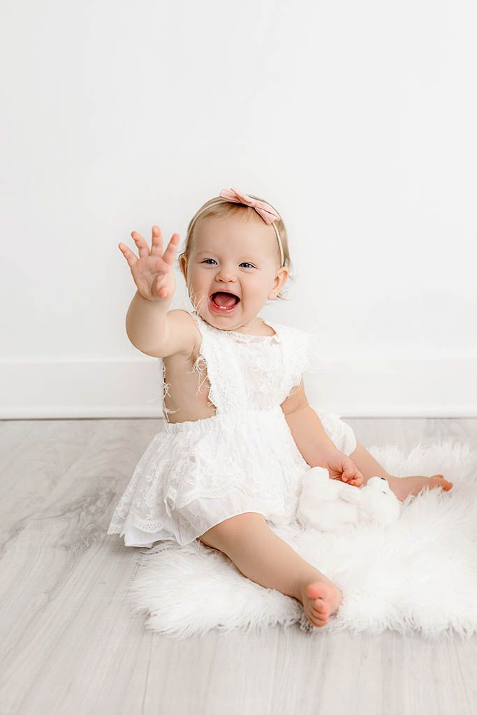Happy baby girl in white lace dress waves at camera during milestone photo session.