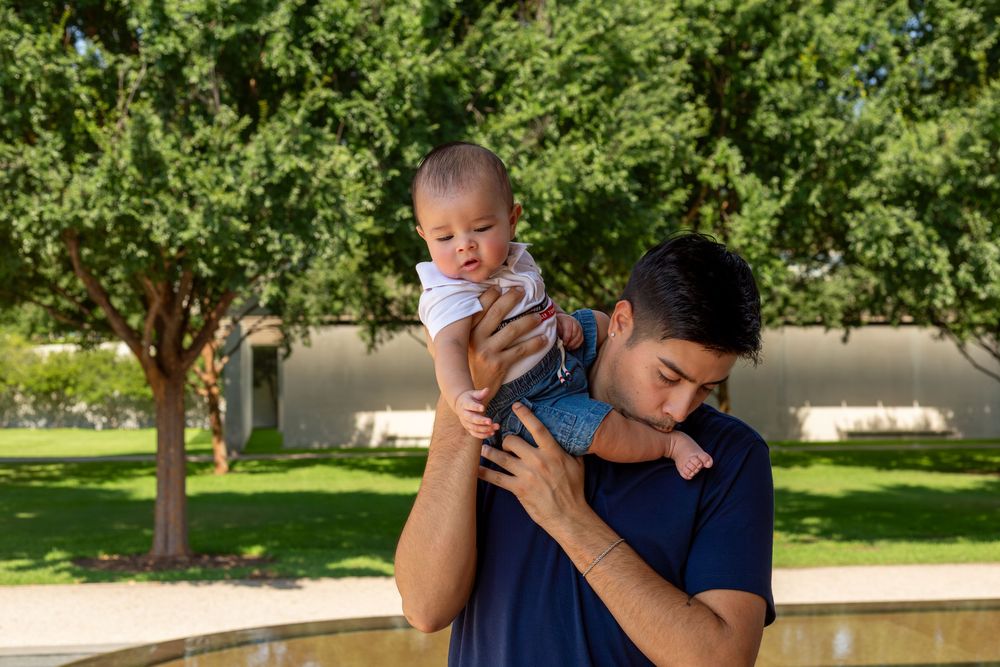 A father lovingly holds his baby up in a sunny park surrounded by lush green trees.