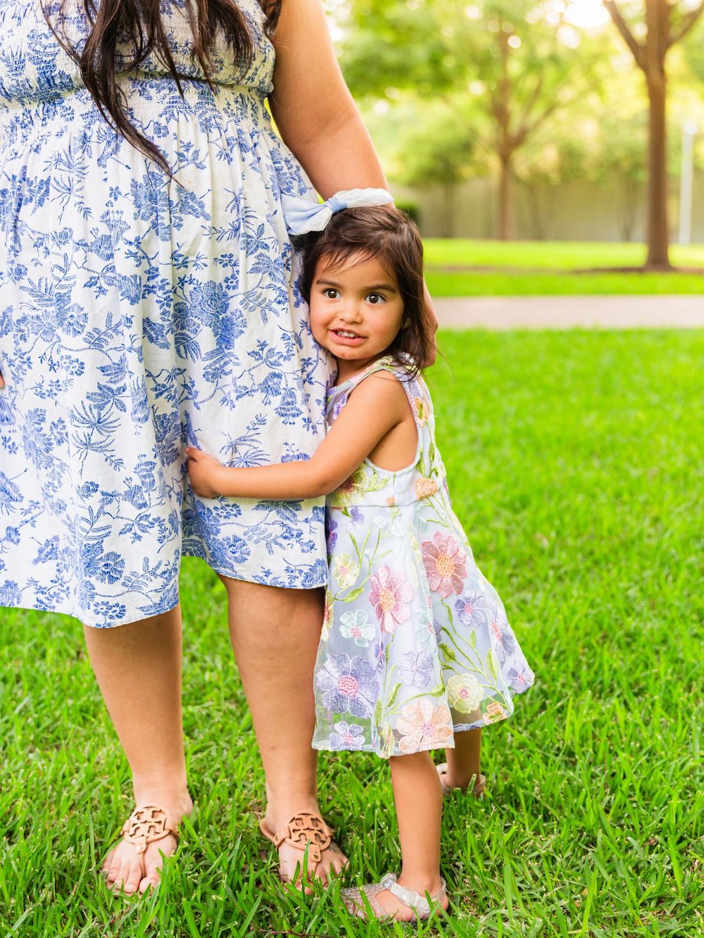 A toddler girl in a floral dress hugs her pregnant mother's belly while standing on green grass outdoors.