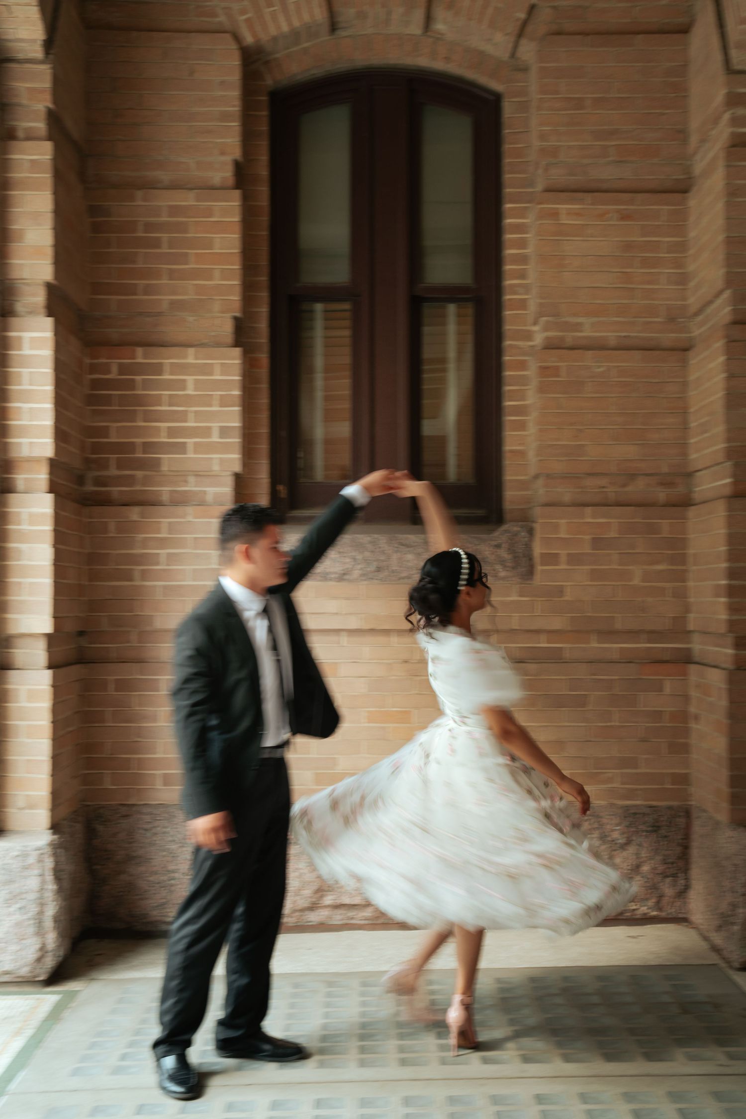 Couple dancing together in front of historic brick building, groom in black suit spinning bride in white dress.